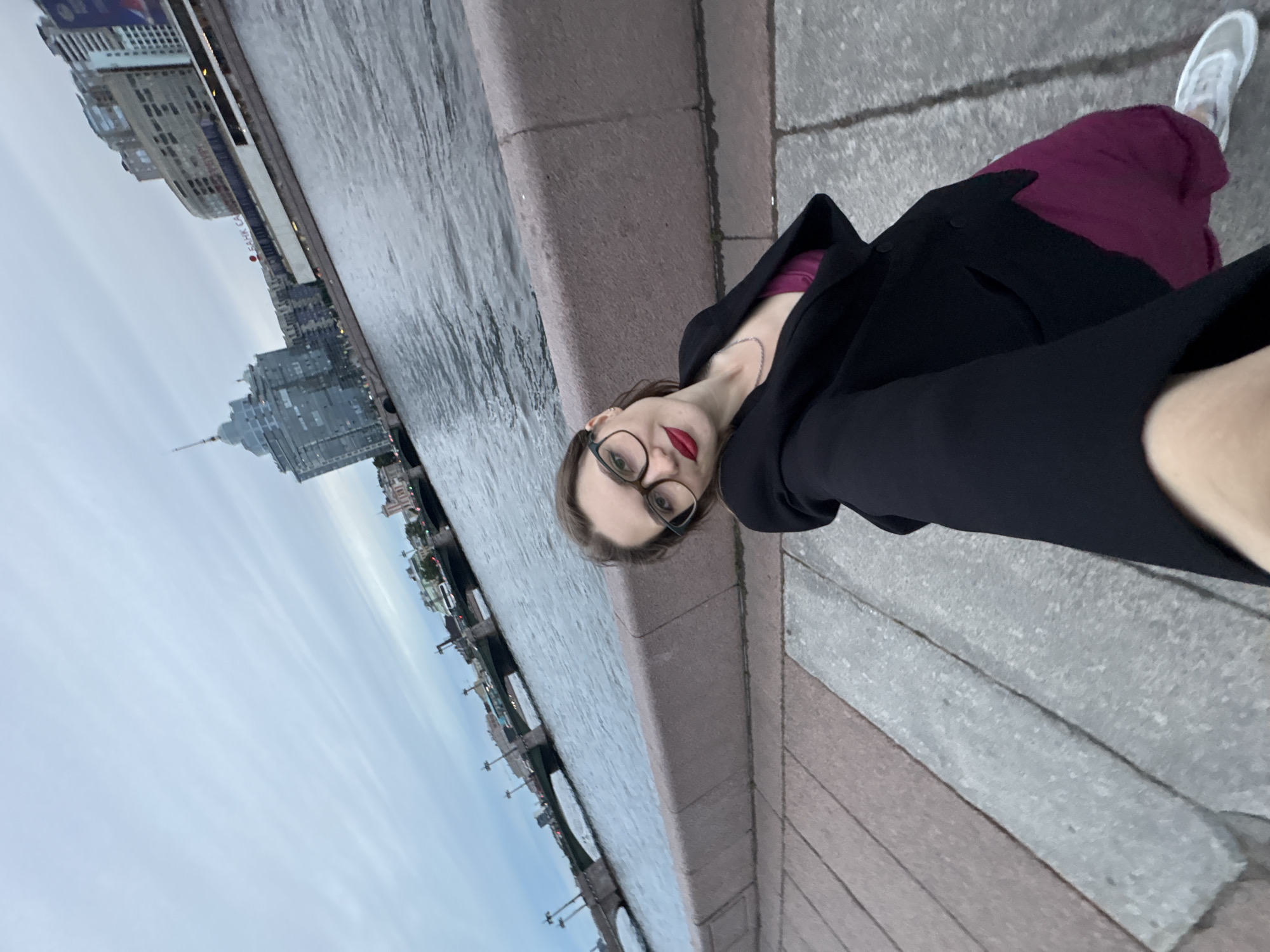 The media file is a rotated, eye-level, full-body selfie taken outdoors in Saint Petersburg, Russia. The subject is a young woman with glasses and red lipstick, wearing a black coat over a purple top and skirt, and white sneakers. She is standing on a stone embankment next to a body of water, possibly a river or canal, with buildings and a bridge visible in the background. The time of day appears to be late afternoon or early evening, with overcast skies. The scene is urban and somewhat muted in color. There is no visible text in the image.