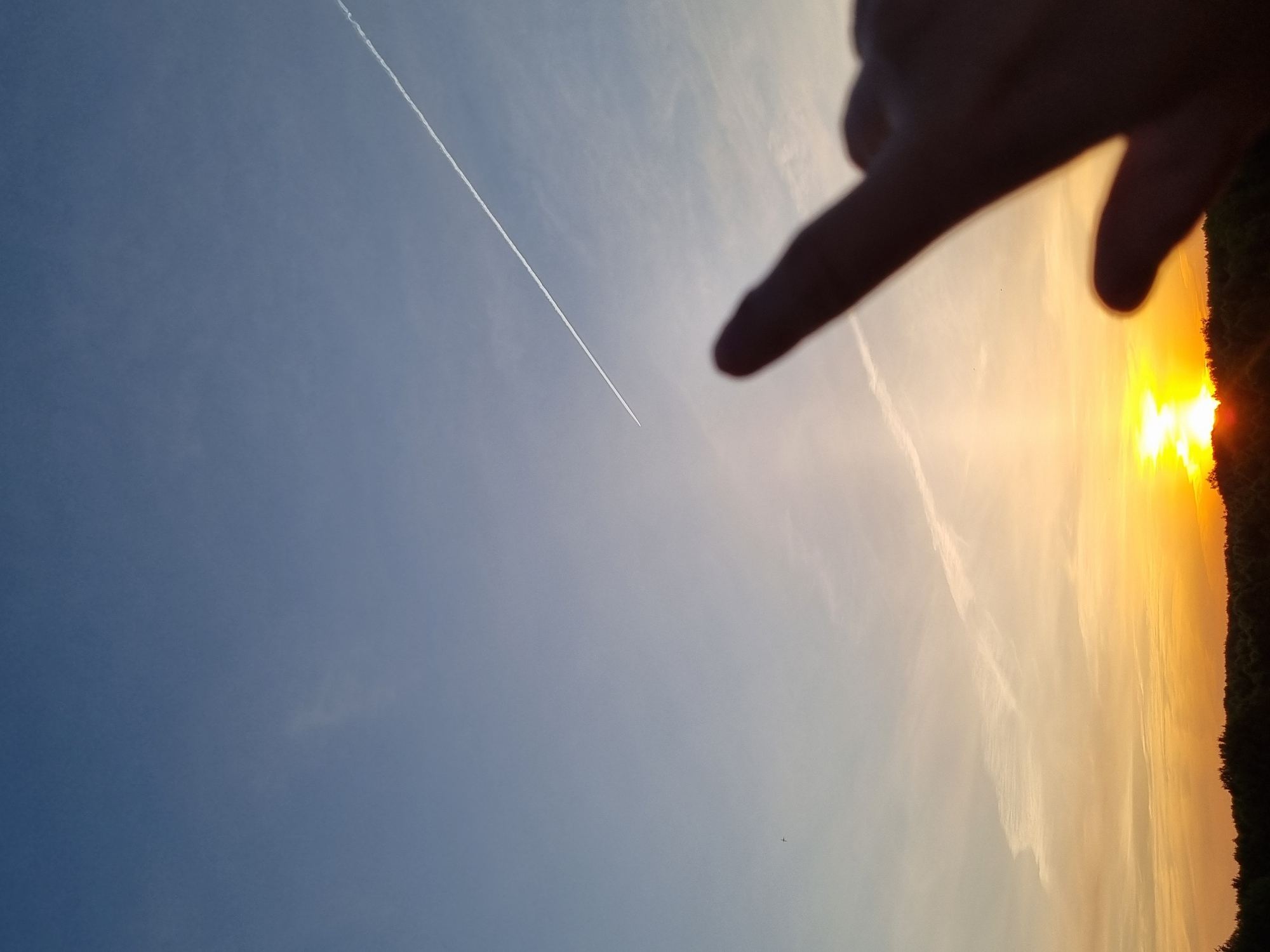 The image captures a sky scene at Kemul, Russia. A contrail stretches across the blue sky. A bright setting sun dominates the right side of the frame, obscured by the silhouette of trees. A hand partially obstructs the view, its fingers blurred in the foreground.
