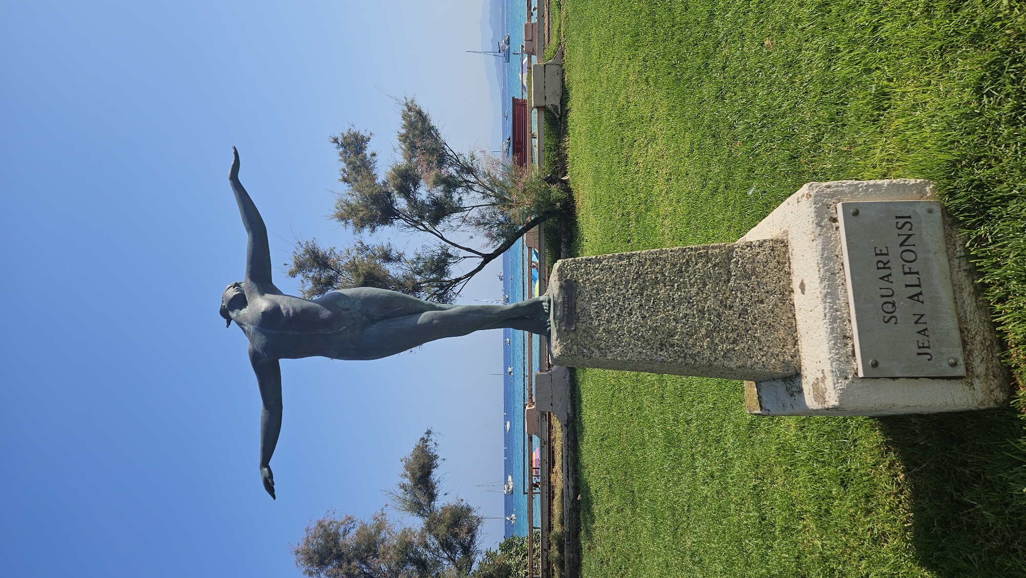 The image shows a bronze statue of a nude female figure with arms outstretched, positioned on a stone pedestal. The statue is located in a grassy park area with a clear blue sky overhead. In the background, there is a body of water with several sailboats, and a beach with lounge chairs and umbrellas. A metal plaque on the base of the pedestal reads "SQUARE JEAN ALFONSI." This suggests the statue is located in Square Jean Alfonsi in L'Île-Rousse, France. The overall scene depicts a serene outdoor setting by the sea.