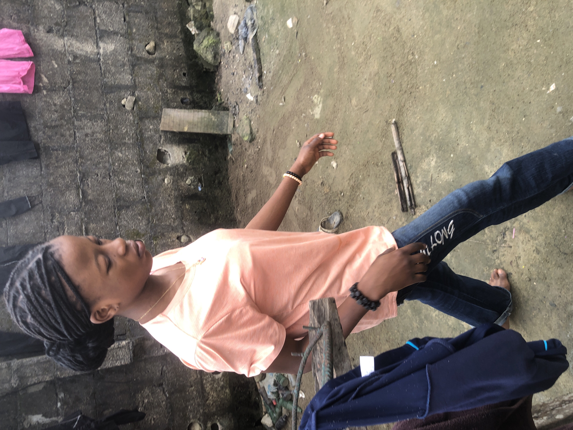 A young Black female with braided hair pulled back in a bun walks to the right, facing away from the camera, in an outdoor environment. She is wearing a light orange t-shirt, dark blue jeans, and beaded bracelets on her left wrist. Her right foot is bare, while her left foot is obscured. A dark blue jacket is draped over a wooden structure to her left. The background consists of a rough, grey concrete wall with some debris and vegetation visible. The ground is a mix of dirt and concrete, with some discarded objects scattered about. The image appears to be taken in Ado, Nigeria, a location suggested by the context provided.