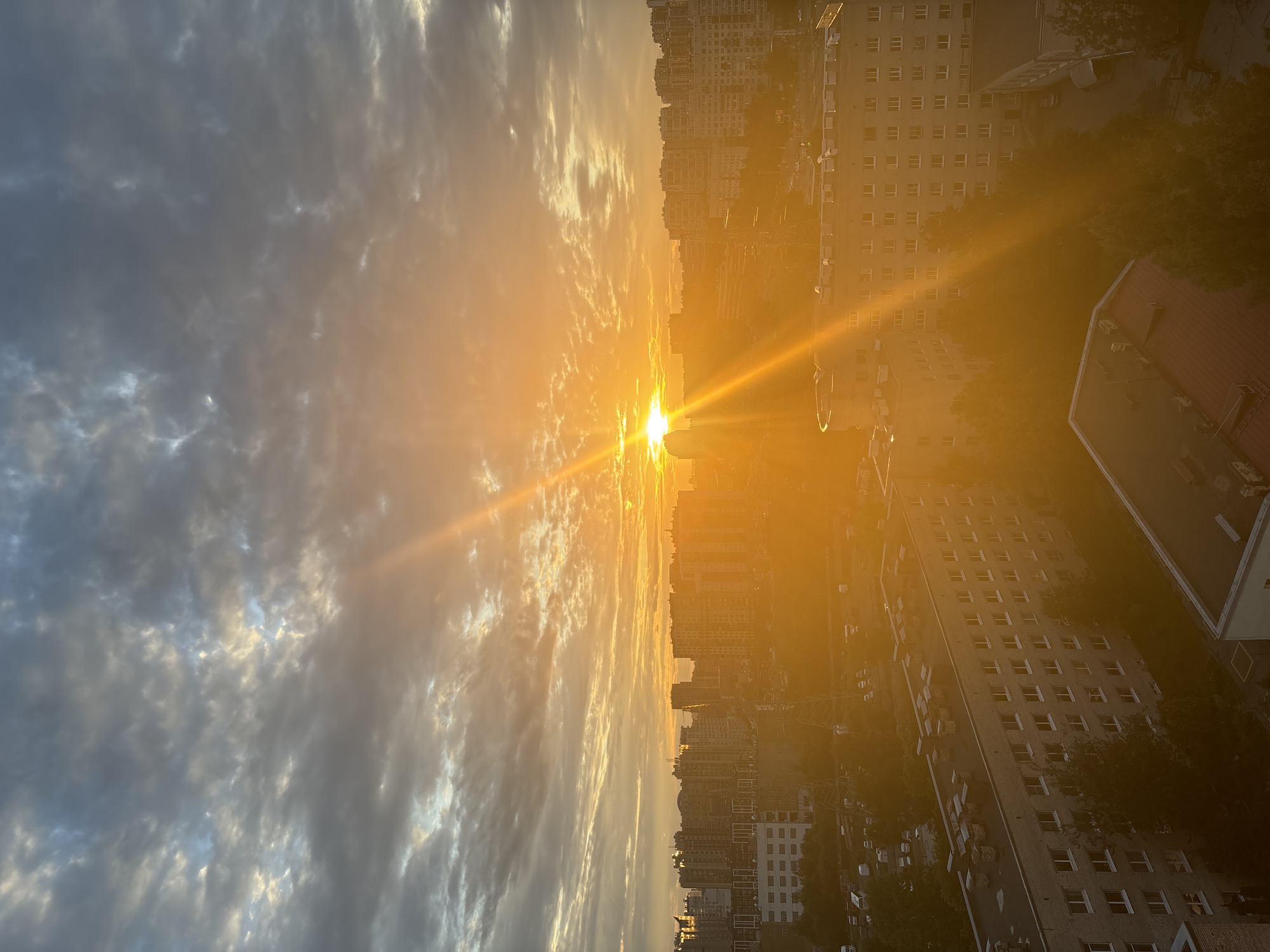 A low-angle, wide shot captures a cityscape under a dramatic sunset. The sky is filled with a mix of dark gray and white clouds, with bright orange and yellow light emanating from the sun, which is positioned slightly left of the center frame. Sunbeams pierce through the clouds, creating a radiant effect. Below the sky, a dense collection of apartment buildings of varying heights stretches across the horizon, predominantly in shades of light gray, white, and beige. The foreground is dominated by closer apartment buildings, some with visible rooftop units and balconies. Trees are scattered throughout the scene, particularly in the middle ground and around the buildings, their forms softened by the sunset's glare. Several vehicles, including vans and cars, are visible in parking areas and on roads amongst the buildings. The overall lighting suggests late afternoon or early evening.
