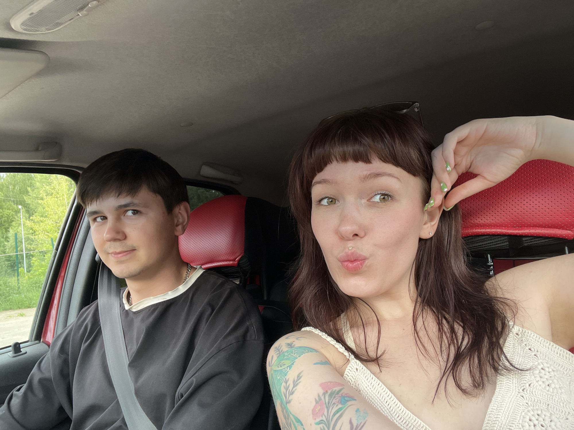 Two individuals are seated in the front of a vehicle, likely a car, which appears to be in a rural or semi-rural area based on the visible greenery and trees outside the window. The person on the left is a young male with dark hair, wearing a black long-sleeved shirt with a light-colored collar and a gray seatbelt. He is looking towards the camera with a slight smile. The person on the right is a young female with dark brown hair, bangs, and light-colored eyes. She is making a kissing face towards the camera and has a lip piercing. She is wearing a light-colored, possibly crocheted, top. Her left arm, visible to the left of the frame, has tattoos of flowers and possibly birds. Her fingernails are painted green with small decorative accents. The interior of the car has red seats visible behind the individuals. The image seems to be a selfie taken from inside the vehicle. The overall lighting suggests it is daytime. The location is not specifically identifiable as Chaikovsky, Russia, from the visual information presented in the image.