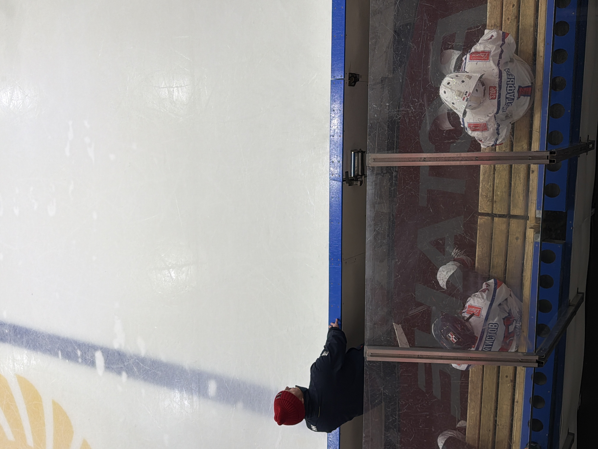 The image depicts an aerial view of an ice hockey rink in Almetyevsk, Russia. To the right of the frame is the players' bench, with two hockey helmets and uniforms visible on the wooden seats. One person, wearing a dark jacket and a red hat, stands at the edge of the bench, near the rink's dasher board. The ice surface occupies the majority of the frame, showing typical skate marks. A blue line and a logo are visible on the ice. The image also captures parts of the rink's surroundings, including the plexiglass barrier and the support structure for the bench.