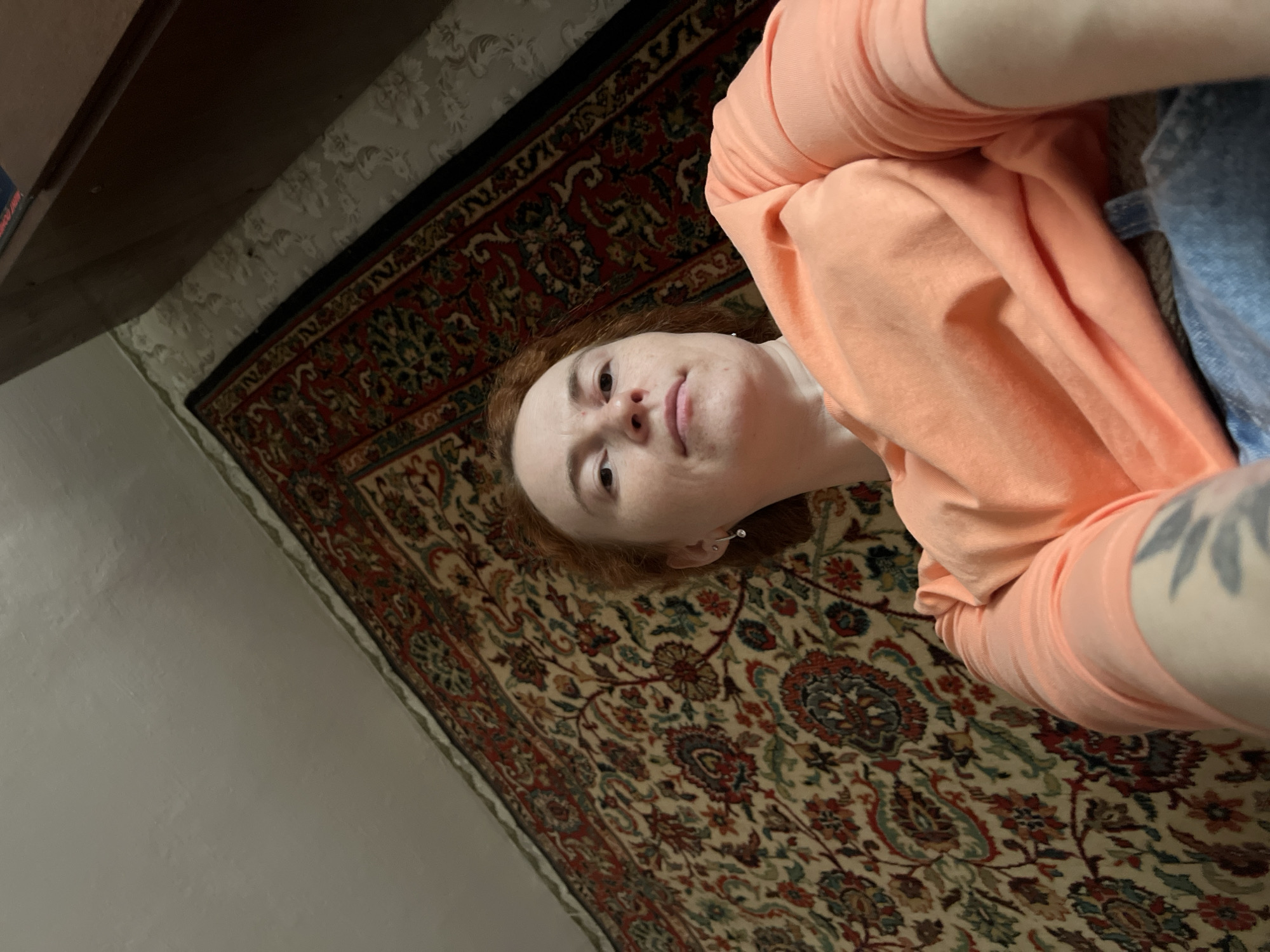 The image presents a woman with reddish-brown hair lying on a patterned rug. She is wearing an orange long-sleeved shirt. Part of a piece of furniture is visible in the upper left corner. The rug features a dense pattern with red, green, and beige tones. The setting is a room with light-colored walls, likely in Chaikovsky, Russia. Details on the scene are limited to what is visible within the frame.