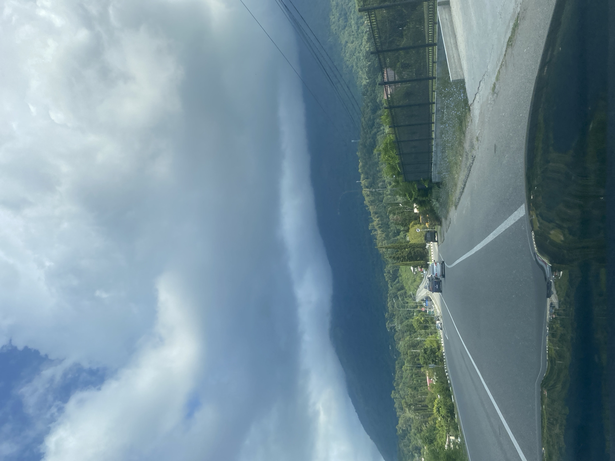 The image depicts a view from a vehicle driving on a paved road in the city of Psakhara, Abkhazia. The road has two lanes with white dividing lines, and there are several vehicles ahead, including a white sedan and a dark-colored SUV. On the right side of the road, there is a black metal fence bordering a property with some buildings visible behind it. To the left of the road, there are trees and houses. In the background, a large, lush green mountain range is shrouded in dark, dramatic clouds. The sky above is a mix of blue and white clouds, with some areas appearing lighter and brighter than others. The overall scene suggests a travel or scenic drive through a mountainous region with some signs of civilization.