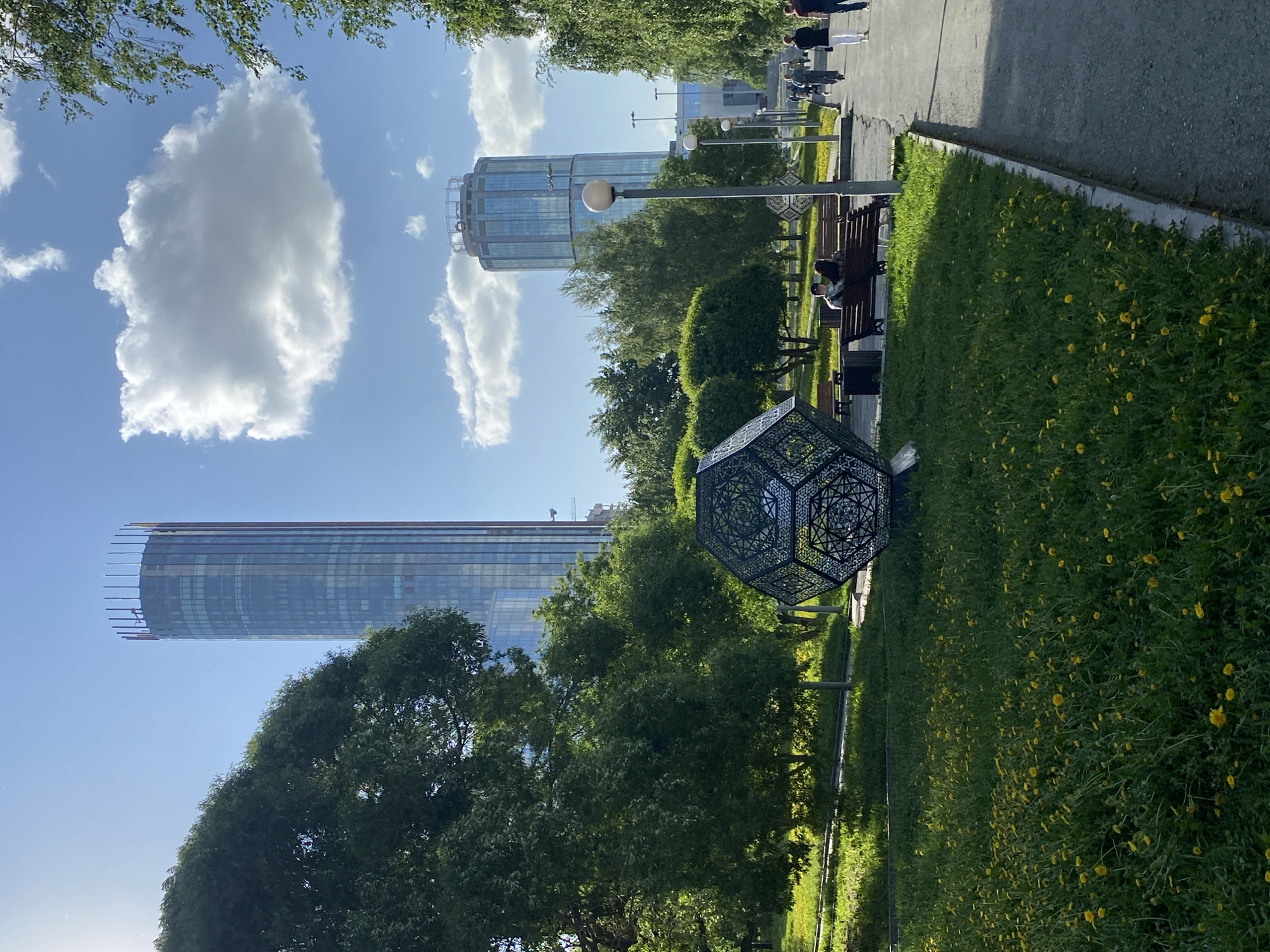 The image captures a scene in Yekaterinburg, Russia, featuring modern architecture alongside a park setting. Two prominent skyscrapers, part of the "Yekaterinburg-City" business district, dominate the background. The foreground reveals a park with lush green grass dotted with small yellow flowers. A geometric, cage-like sculpture stands prominently near the edge of the park. A paved path runs along the edge of the park, with benches placed along it. People are sitting on one of the benches. Overhead street lamps are visible. The sky is blue with scattered cumulus clouds.