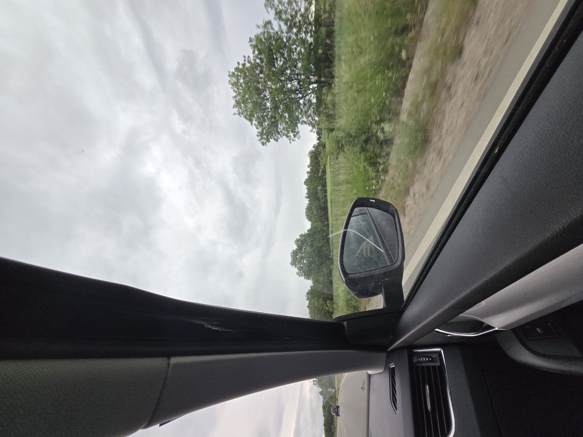 The image shows a view from inside a car driving on a road in an unknown location. The sky is overcast with grey clouds. A tree and grassy vegetation line the roadside. The car's side mirror is visible, and it appears to be cracked. The interior of the car, including the dashboard and air vents, is also visible. Another vehicle is seen in the distance.