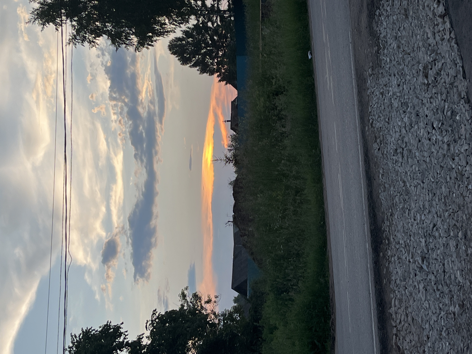 The image depicts a rural landscape at dusk, likely in Ludorvay, Russia. The sky is filled with a mix of clouds, with streaks of orange and yellow from the setting sun visible on the horizon. Power lines stretch across the upper portion of the frame. In the middle ground, there are several structures, including a dark-roofed building and a light-colored fence. Dense green vegetation covers the lower middle ground, with a pile of dark material to the left of the building. A paved road runs across the foreground, bordered by a section of loose gravel. Trees frame the left and right sides of the image.