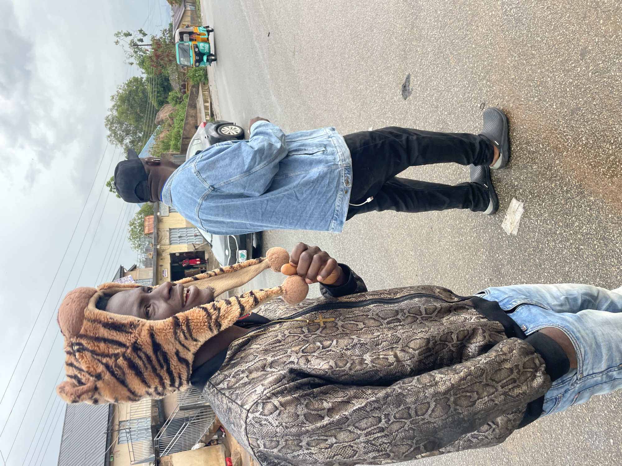 Two men stand on an asphalt street in Jos, Nigeria, with buildings and a "keke" (tricycle taxi) in the background under an overcast sky. The man in the foreground wears a tiger-striped hat, a snake-print jacket, and jeans, holding a pair of small, round, brown objects, possibly gourds or wooden balls, in his right hand. The man in the background wears a denim jacket, black pants, and a black cap, standing with his back to the camera. A silver-colored car is parked nearby.