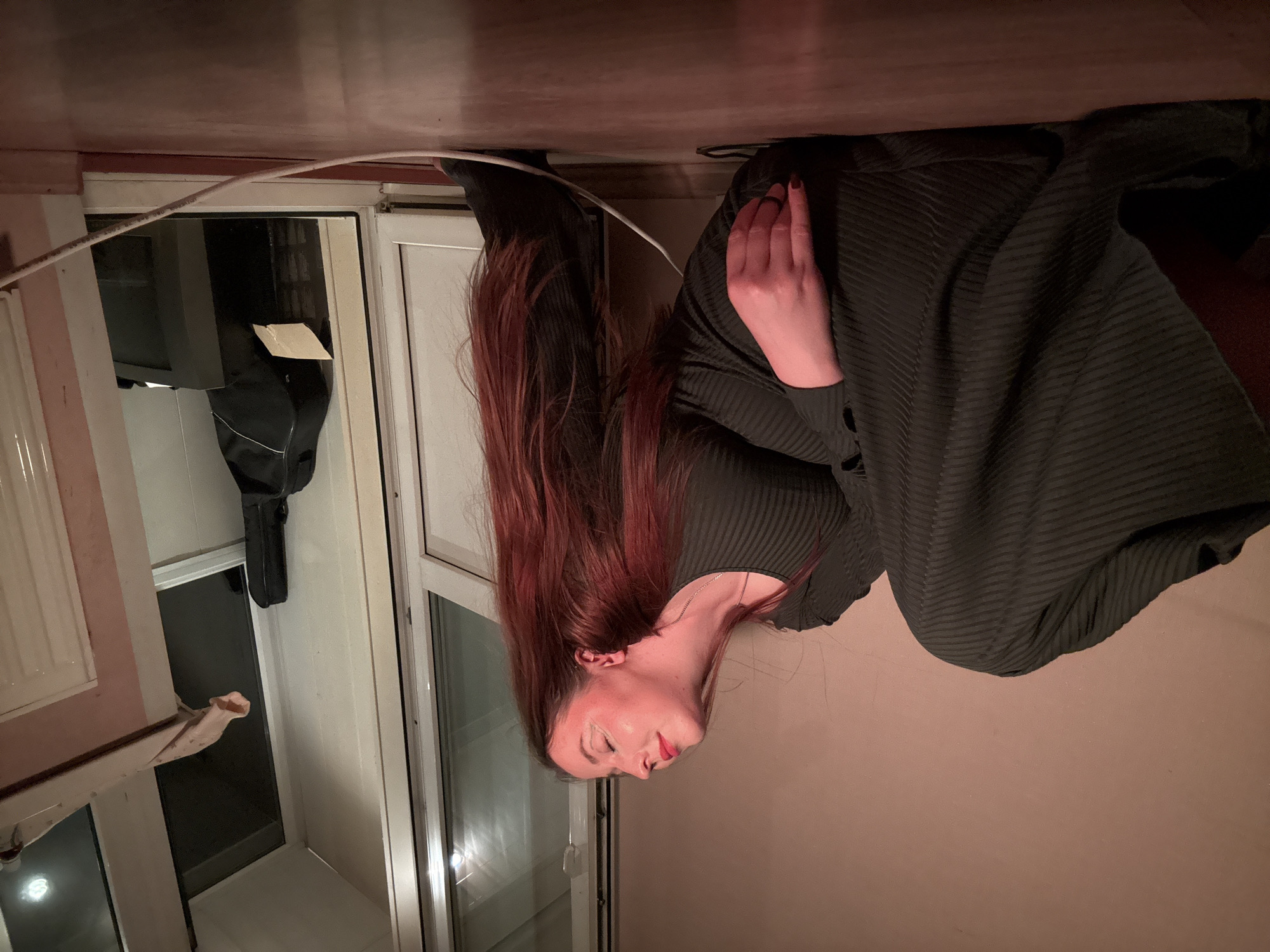 A woman with long reddish-brown hair is seen in an upside-down view, positioned close to a window. She is wearing a dark green ribbed shirt or dress and is standing indoors. A white cable runs along the top of the window frame. Outside the window, a guitar case hangs from a door. The location is unknown and has not been provided.