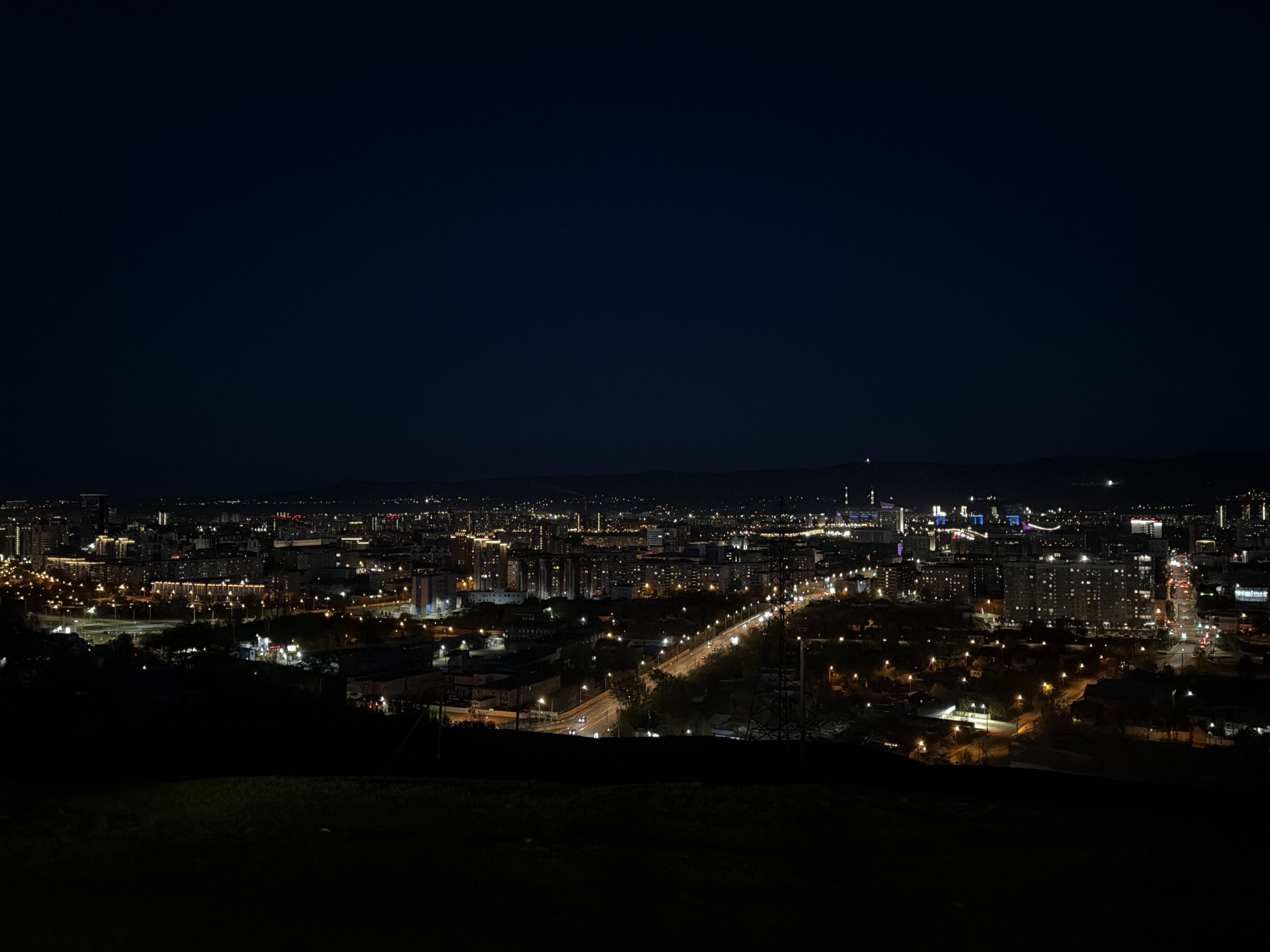 The image shows a night view of Krasnoyarsk, Russia, from an elevated perspective. A dark foreground of grass and trees leads to the cityscape, illuminated by numerous streetlights and building lights. A major road cuts through the center of the frame, with headlights visible on moving cars. In the distance, the city skyline features taller buildings and structures. The sky is a deep, dark blue. There are no people visible in the image.