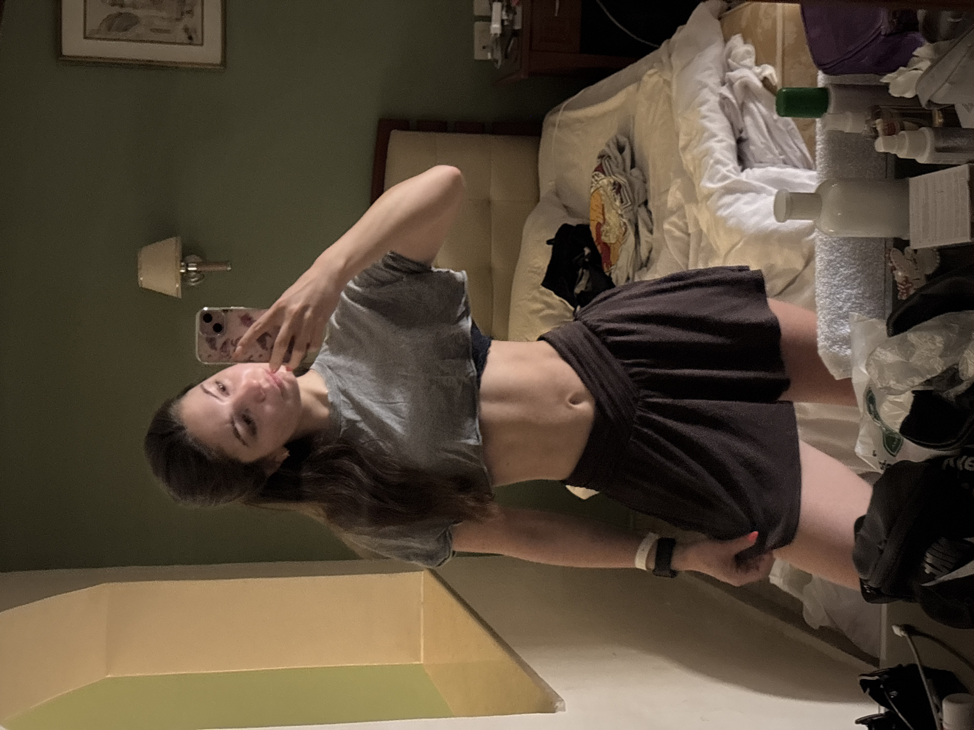 A woman is taking a selfie in a room. The room contains a bed with bedding, a nightstand, and various items on the nightstand, including toiletries. The wall is painted a dark color, with a picture hanging on it. The location is unknown.