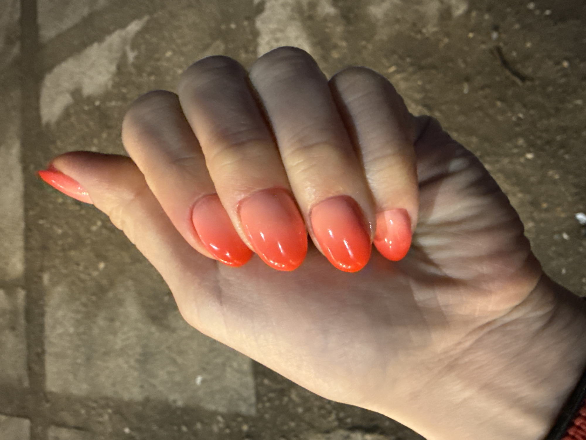 The image presents a close-up view of a hand, likely female, showcasing an ombre nail art design. The hand is positioned with fingers slightly curled, drawing attention to the nails against a dark, textured background.
The nails are uniformly shaped, appearing oval or almond. They feature a glossy, two-tone ombre effect, transitioning from a lighter, soft peach or pink hue at the cuticle to a vivid, bright orange-red at the tips. The skin of the hand appears smooth, with natural creases and lines visible on the knuckles and palm, indicating normal skin texture.
The scene is dimly lit, with shadows cast across the fingers and the background, suggesting that the photograph was taken at night or in a low-light indoor setting. The background is a rough, grayish surface, consistent with asphalt, concrete, or stone pavers. No specific activity is taking place beyond the presentation of the hand and nails. There is no visible text in the image. The location context for this image is Chaikovsky, Russia.