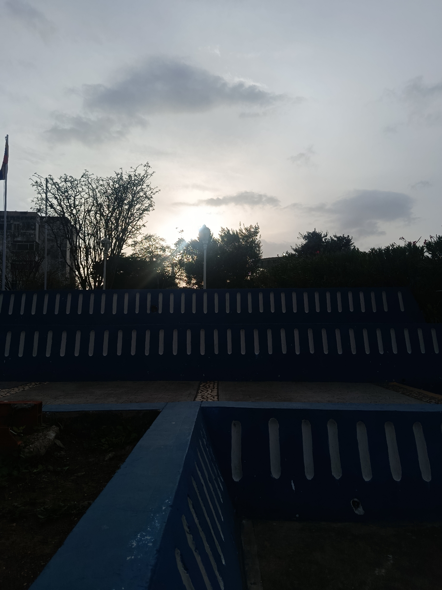 The image depicts a twilight scene at a park or plaza in Juan Griego, Venezuela. In the foreground, a blue, two-tiered concrete structure with vertical rectangular cutouts dominates the view. Beyond the structure, a paved area is visible, bordered by trees and foliage. In the background, partially obscured by the foliage, is a building. A flag, possibly Venezuelan, is on a pole to the left of the building. The sky is overcast with the sun low on the horizon, providing a diffused, back-lit view. The scene appears still and deserted.