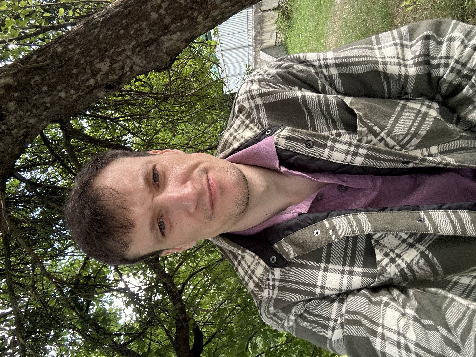 A man is taking a selfie outdoors, with a large tree behind him. He is wearing a green and white plaid shirt over a pink polo shirt. The man is looking directly at the camera with a neutral expression. He has short brown hair and light-colored eyes. The background is out of focus but appears to be a grassy area with a white fence and some vegetation. The lighting suggests it is daytime. The location is Makiv, Ukraine.