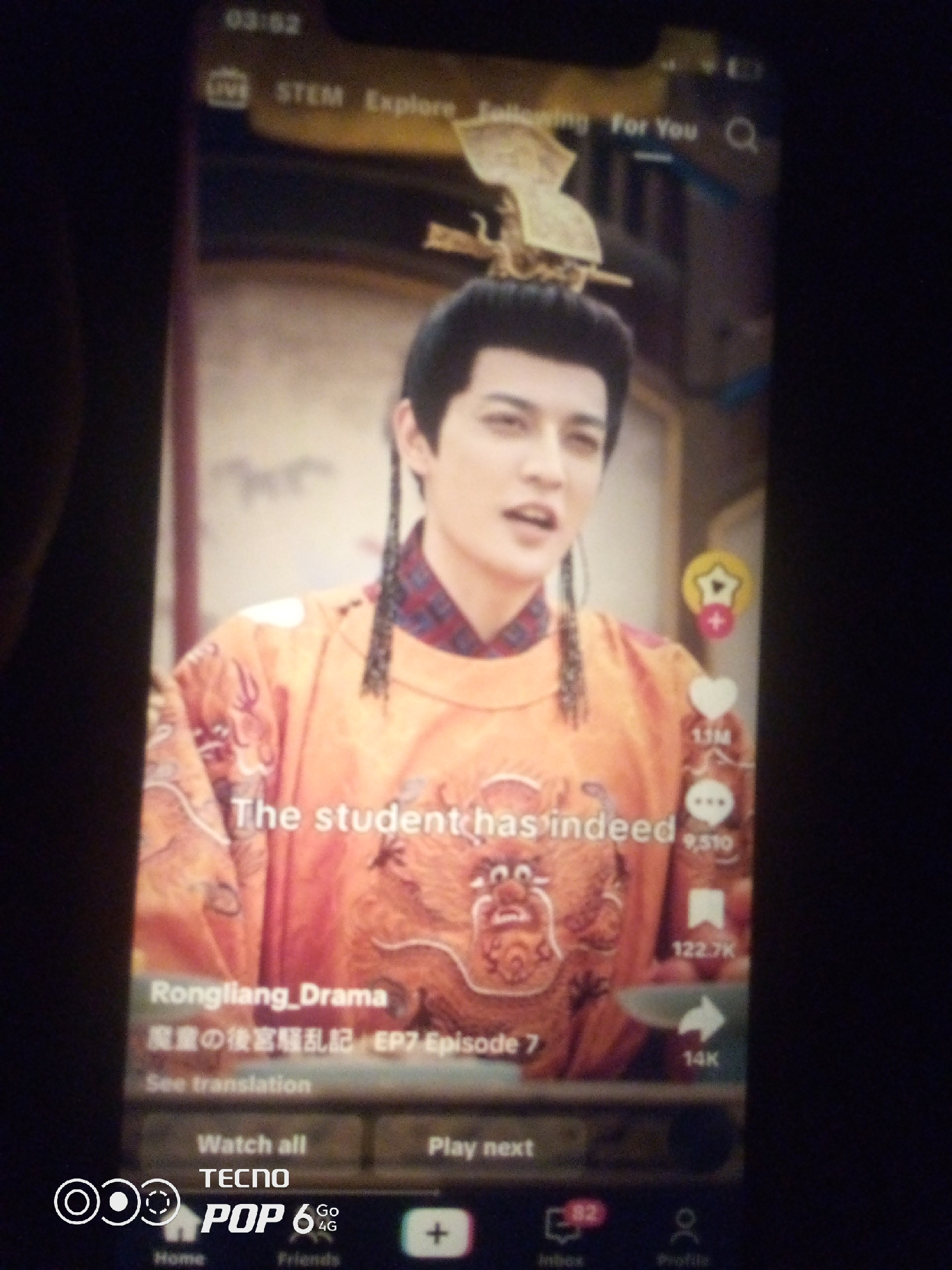 The image shows a TikTok video playing on a smartphone. The video features a person dressed in traditional Chinese attire with elaborate hair and makeup, an orange robe adorned with a dragon motif. The text overlay on the video reads "The student has indeed" and "Rongliang_Drama," followed by Chinese characters and "EP7 Episode 7." There are also engagement icons like a heart with "1.1M" likes and a comment icon with "9,510" comments. At the bottom of the screen, a Tecno Pop 6 4G interface is visible with navigation icons for "Home," "Friends," "Inbox," and "Profile." The smartphone is positioned in what appears to be a dimly lit indoor environment, possibly a home or a public space in Howayi, Nigeria, although there are no specific visual cues to confirm the exact location within the city. The overall image quality is low due to the photographic conditions.