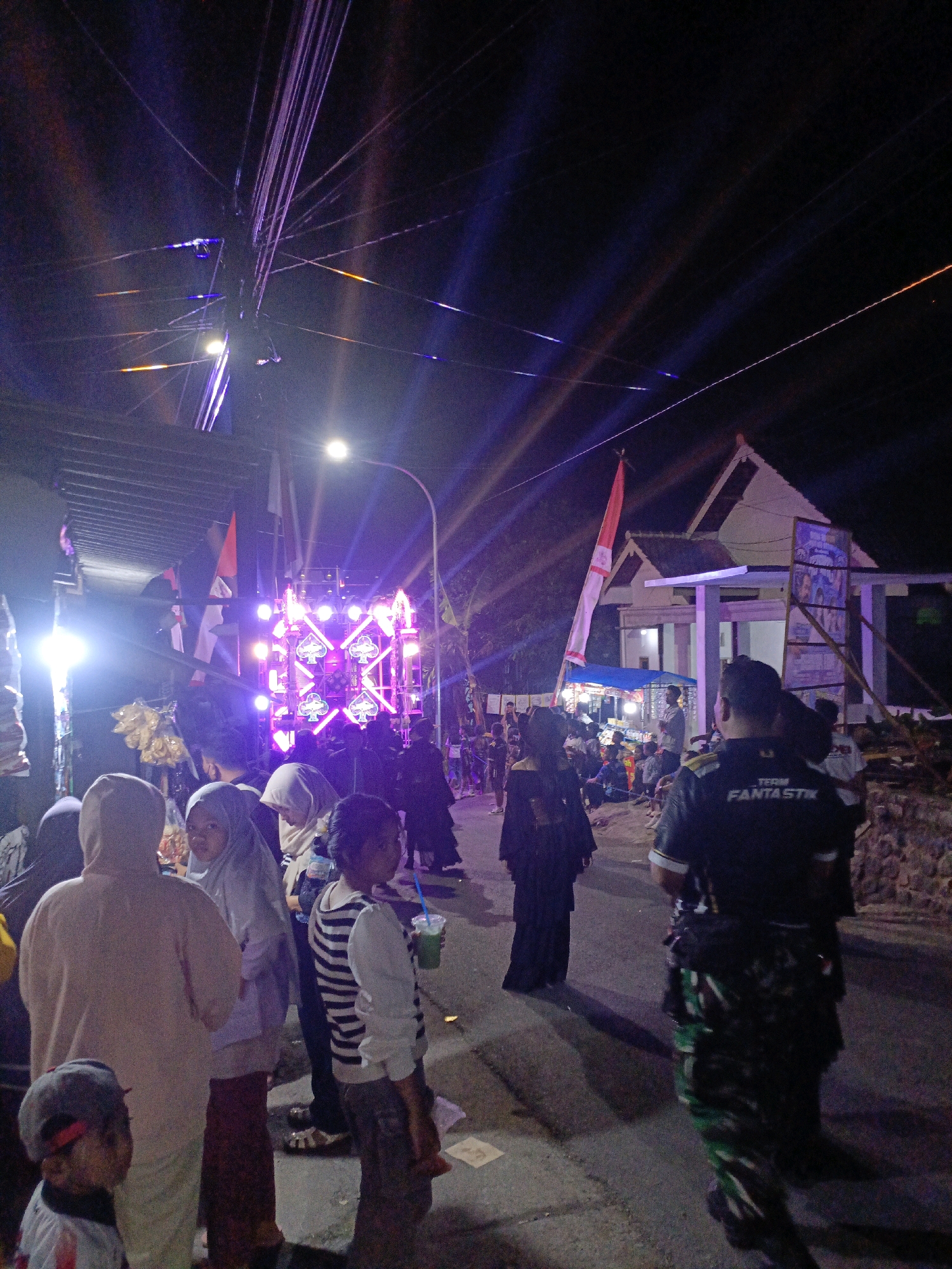 The image depicts a nighttime scene in Pager, Indonesia, likely during a local festival or event. A large, illuminated sound system is a central feature, with bright purple and pink lights creating a vibrant atmosphere. Several people are visible, including children and adults, many of whom appear to be observing the spectacle or socializing. Some individuals are wearing traditional Indonesian attire, such as hijabs. A man in the foreground wears a dark shirt with the words "TEAM FANTASTIK" printed on the back, suggesting he might be part of an organizing group or entertainment crew. The background shows buildings and streetlights, indicating an urban or semi-urban setting. The overall impression is one of a lively community gathering.
