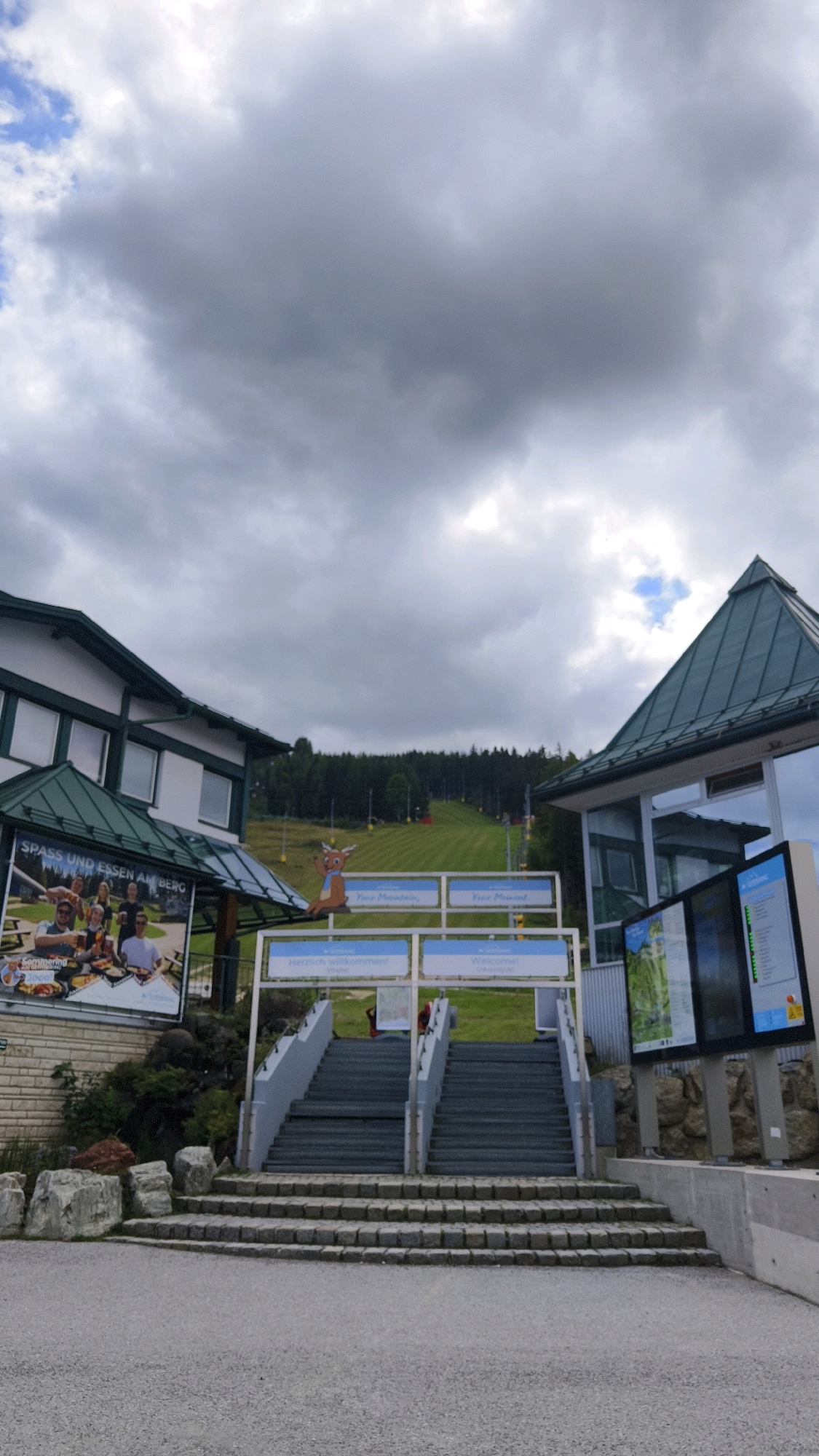 The image captures an outdoor scene at the base of a ski slope in Spital am Semmering, Austria. The sky is overcast with dramatic, grey clouds. In the foreground, a paved area leads to a set of wide, grey concrete stairs that ascend upwards. To the left of the stairs is a building with a green roof and white walls, featuring a large advertisement poster. The poster depicts people enjoying food and drinks, with text including "SPASS UND ESSEN AM BERG" (Fun and Food on the Mountain). A small, cartoon-like deer figure is seated on the railing above the stairs.
To the right of the stairs is another building with a prominent green, pointed roof and large glass windows that reflect the cloudy sky. Mounted on the exterior of this building are digital display screens showing maps and information.
The ski slope itself rises behind the buildings and stairs, with neatly mown green grass and visible ski lifts. A dense forest borders the top of the slope. The overall impression is a functional, tourist-oriented location, likely for summer activities given the green slope.