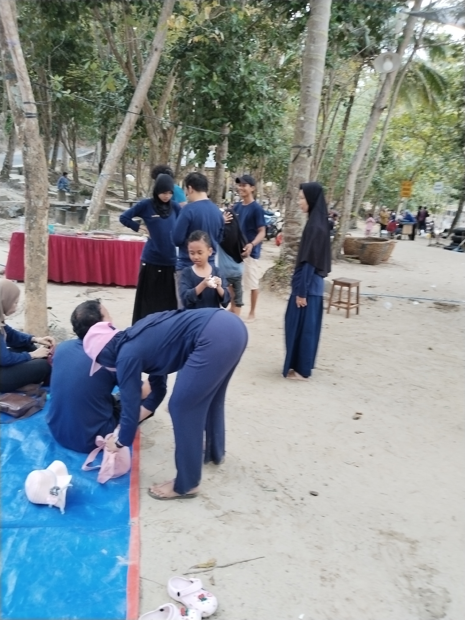 The image captures a beach scene in Prigi, Indonesia, with several individuals present. In the foreground, a person in a navy blue outfit is bending over, seemingly interacting with a pink bag and a white sandal on a blue tarp. Another person in a similar navy blue outfit is seated on the tarp to the left, with a brown bag nearby. Behind them, a young girl in a black shirt is looking down at something in her hands. Further back, a woman in a black hijab and navy blue dress is standing, and to her left, two young men in blue shirts are also visible, with one wearing a black cap. The environment is sandy, with trees and foliage forming a backdrop. In the distance, there are more people, some sitting and others walking, and a red-covered table. A wooden stool is placed on the sand near the woman in the black hijab.