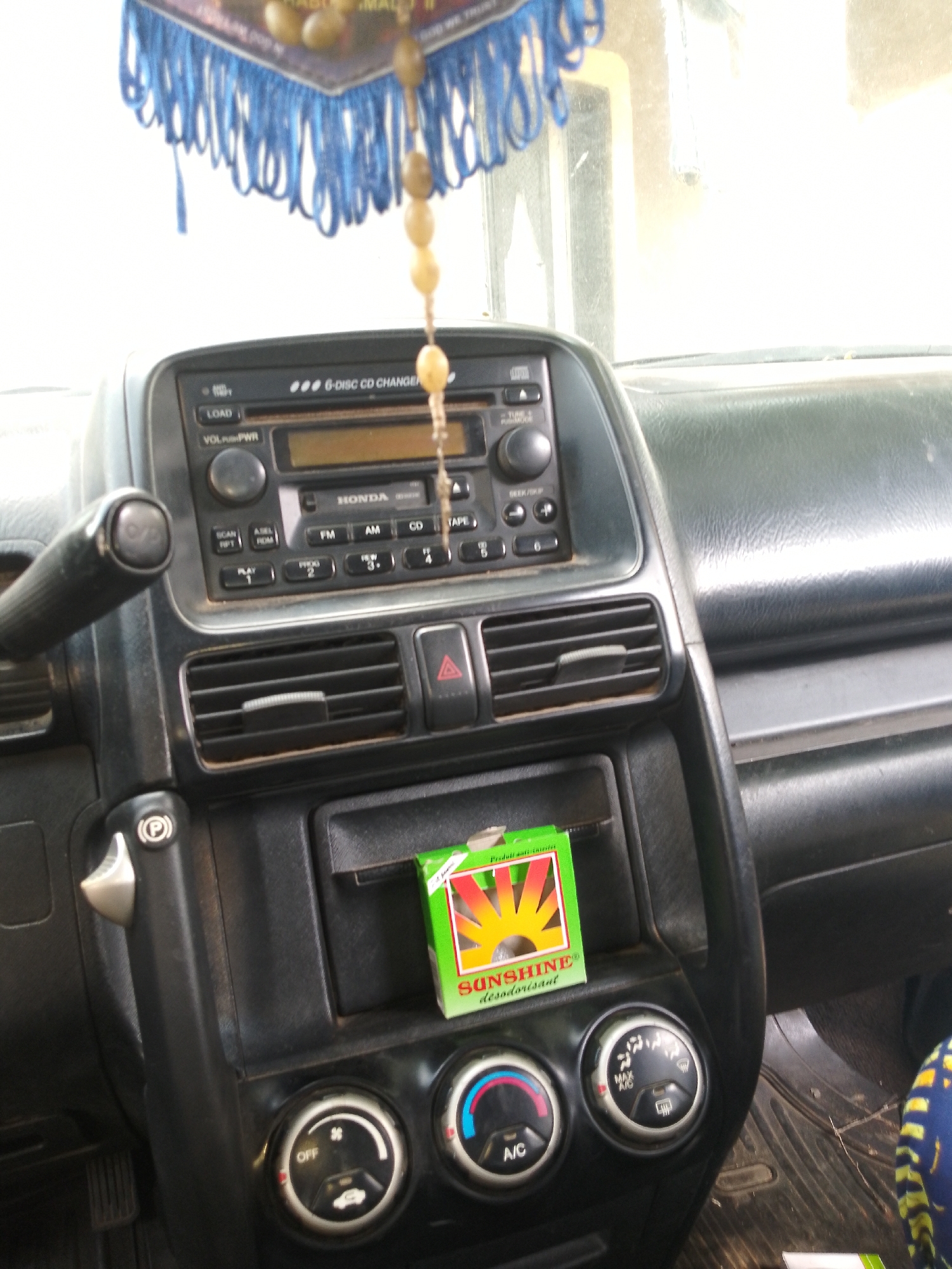The image shows the interior of a car, specifically the dashboard and the area around the center console. A car radio with a CD changer is visible. To the right of the radio, there are air vents and controls for the climate system. A small green box with a sun graphic, labeled "SUNSHINE désodorisant," is placed on the console below the air vents. A handbrake lever is also visible on the left side of the console. The car appears to be parked, as indicated by the engaged parking brake symbol on the lever. The overall environment suggests an indoor or covered setting, possibly a garage or a parking lot, with natural light coming from the front. The image does not contain any information about the city of Serti, Nigeria, or any people.