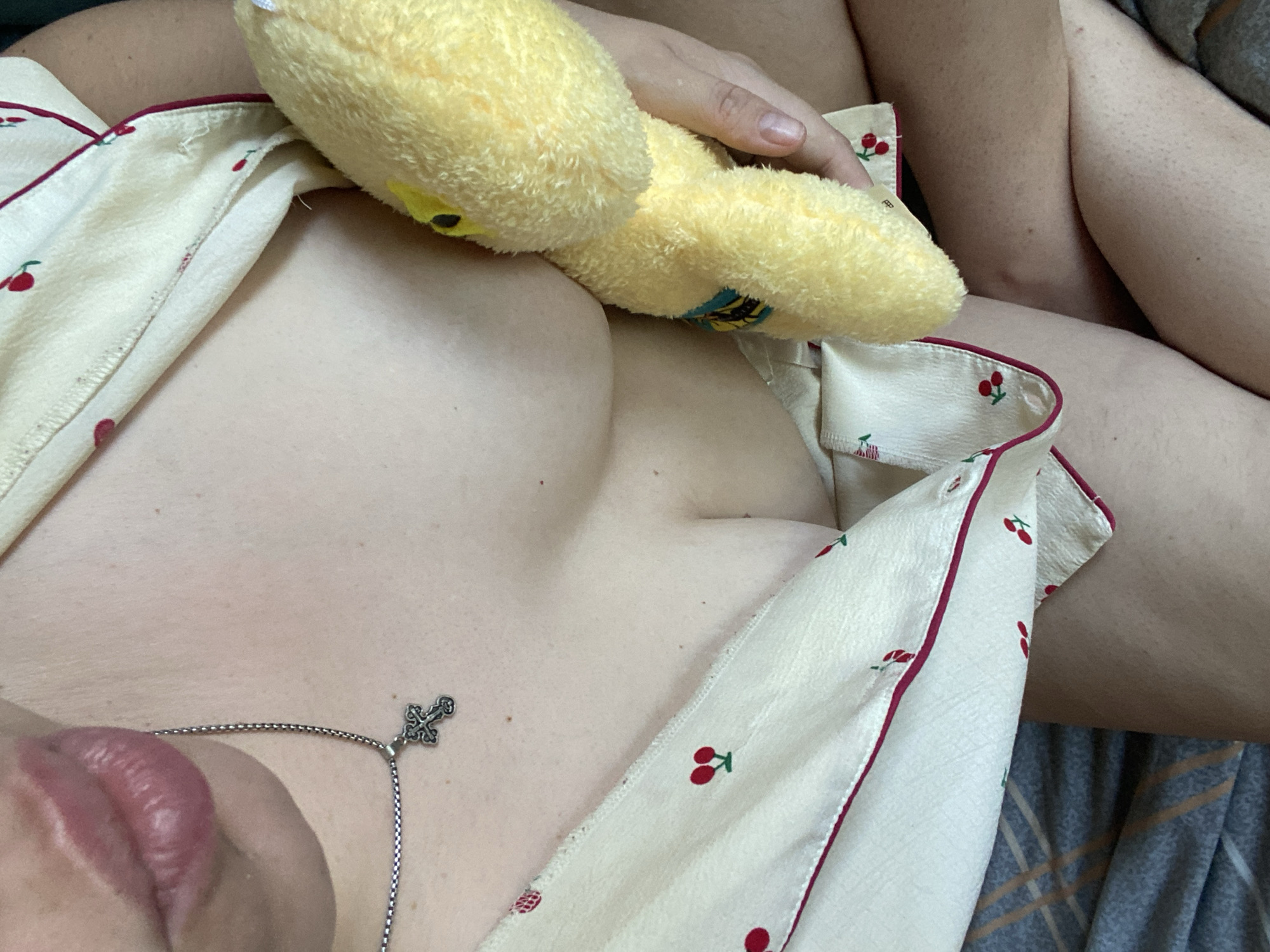The image shows a close-up, cropped view of a person's upper body and legs. The person is wearing a light-colored pajama top with a cherry print and red trim. A silver necklace with a cross pendant is visible on the person's chest. The person is holding a yellow stuffed toy. The background consists of bedding, with a patterned sheet visible. The scene appears to be indoors, likely in a bedroom. The lighting is soft, suggesting natural light or indoor lighting. The exact location within the city of Unknown, Unknown cannot be determined from the provided image.