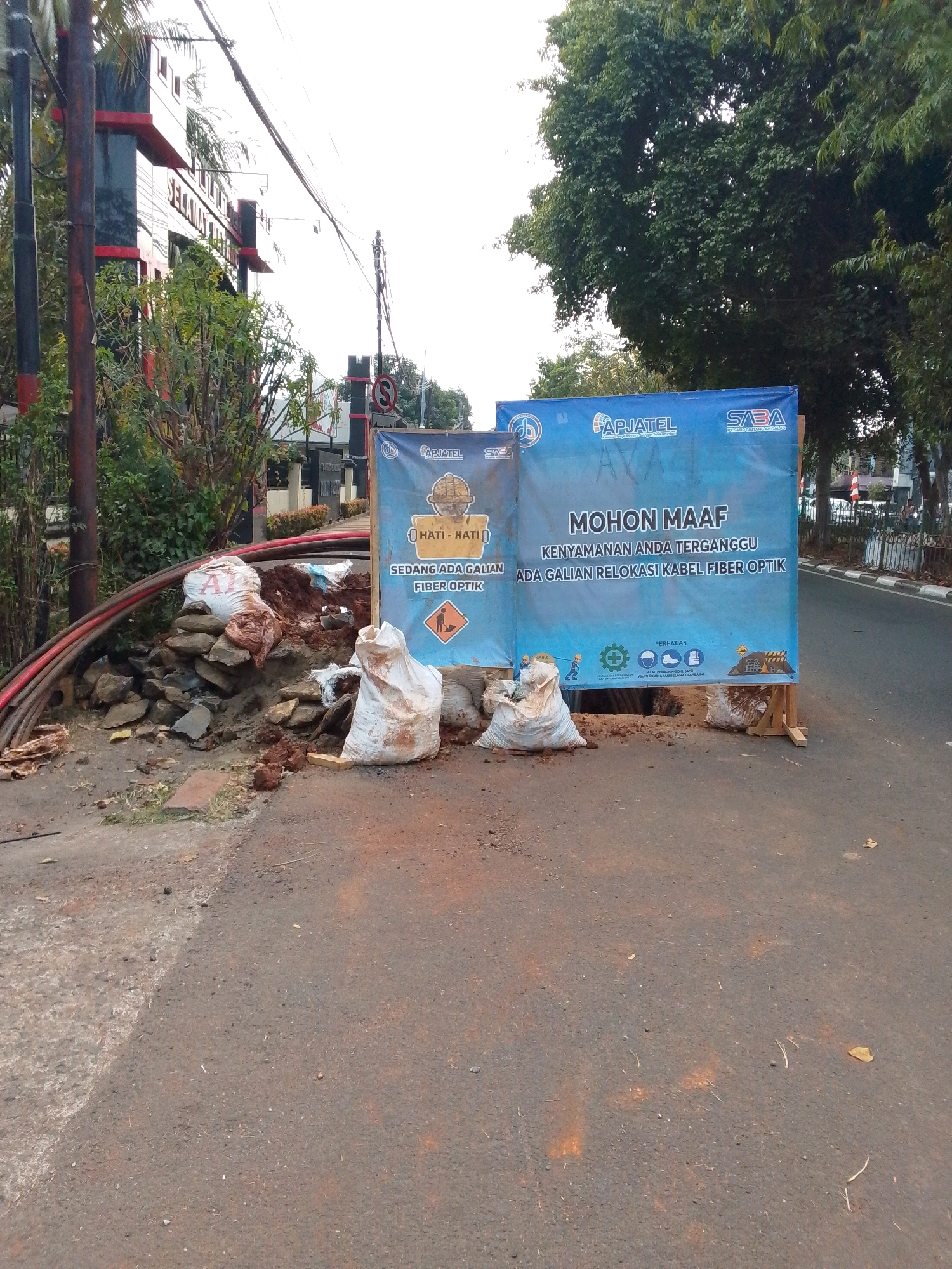 The image depicts a street scene in East Jakarta, Indonesia, with ongoing fiber optic cable relocation work. A large blue banner is prominently displayed, conveying a message of apology for the inconvenience caused by the excavation. The banner reads in Indonesian: "MOHON MAAF KENYAMANAN ANDA TERGANGGU ADA GALIAN RELOKASI KABEL FIBER OPTIK," which translates to "WE APOLOGIZE FOR THE INCONVENIENCE, THERE IS A FIBER OPTIC CABLE RELOCATION EXCAVATION."
The excavation site is located on the side of the road, marked by piles of dirt and rocks, and several white sacks filled with soil. Rolls of thick, colored cables (red and black) are visible near the excavation, suggesting they are being worked on or have been removed. The area is cordoned off, with safety signs indicating potential hazards, including a warning symbol for excavation and a helmet icon.
In the background, a modern building with red and black accents is partially visible, along with lush green trees lining the street. The sky is overcast, and the overall lighting suggests it is daytime. The presence of traffic signs, including a circular sign with a red border and an 'S' symbol, indicates this is a public road. The general environment is a typical urban street with infrastructure work underway. There are no people clearly visible in the image actively working on the site, but the presence of the banner and the ongoing work strongly imply human activity.