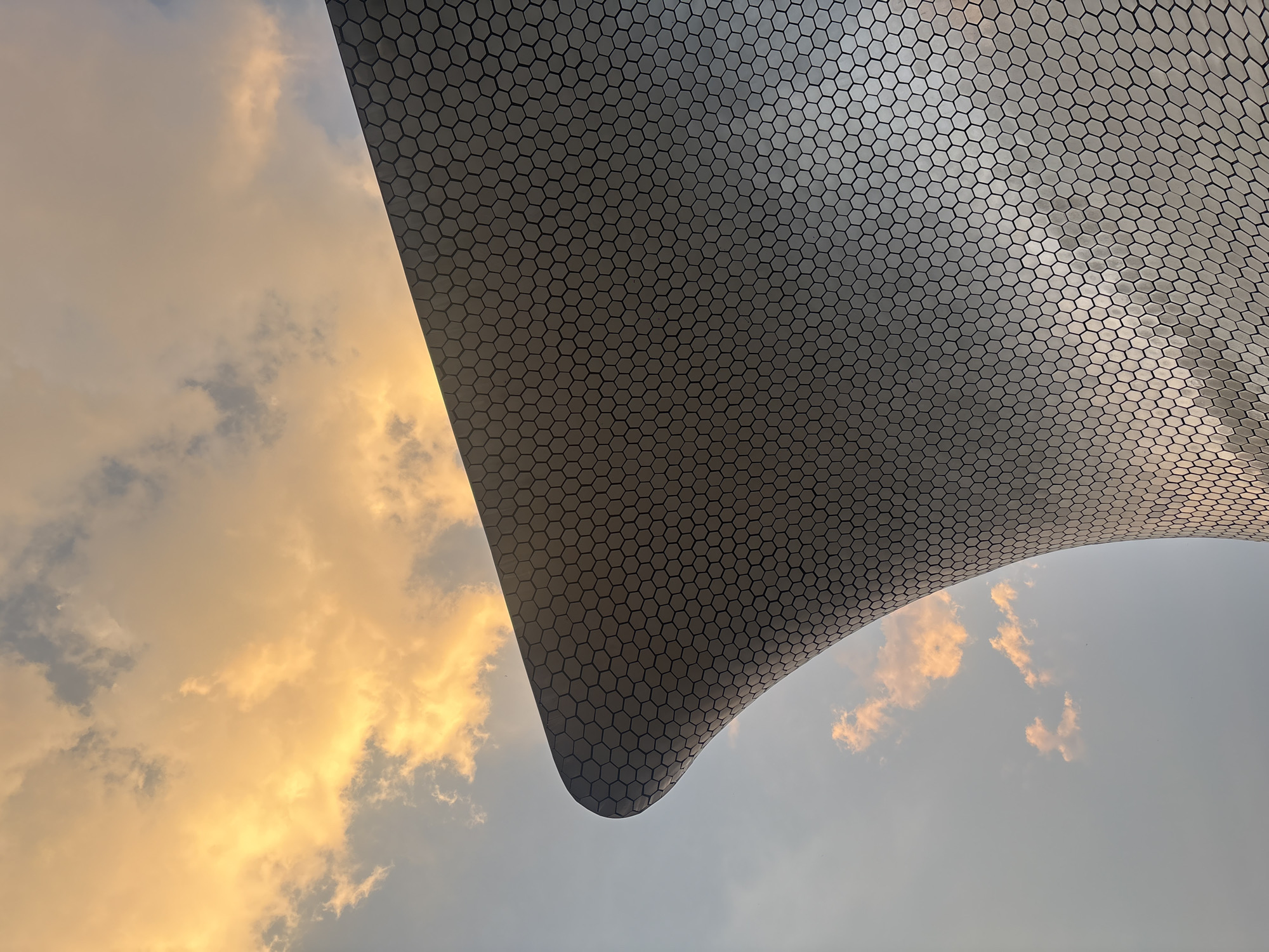 The image captures a partial view of the Soumaya Museum in Mexico City, Mexico. The museum's distinctive architecture, characterized by an asymmetrical, curving form covered in hexagonal aluminum panels, is prominently displayed. The sky serves as a backdrop, with warm, golden hues near the horizon contrasting with the cooler tones higher up. Clouds are scattered throughout the frame, contributing to the dynamic composition. The perspective is from below, looking upward towards the building's exterior.