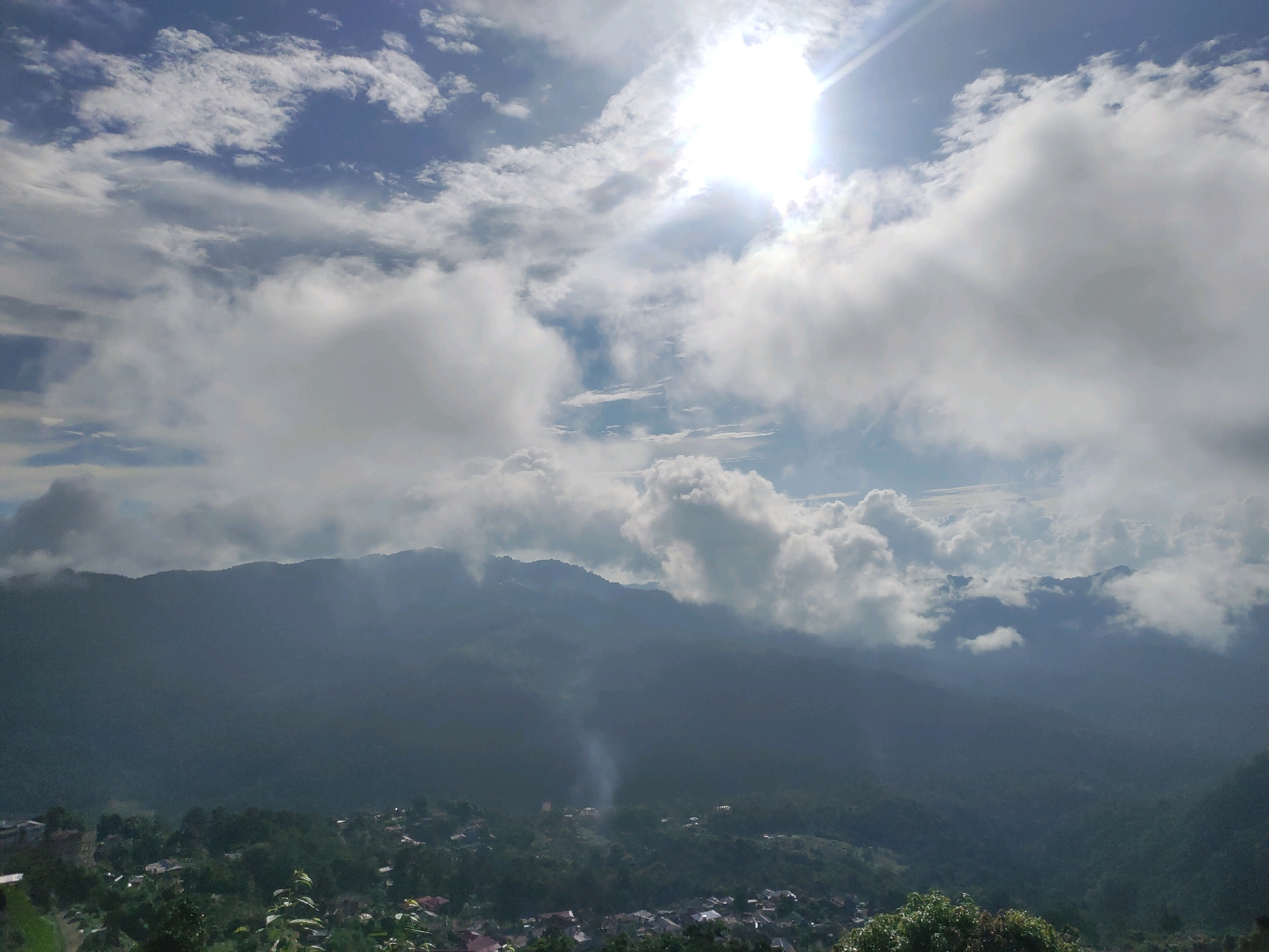 The image depicts a panoramic view of a mountainous landscape under a bright, partly cloudy sky. The sun is prominently visible in the upper right quadrant, casting a strong glare and lens flare. Cumulus clouds are scattered across the sky, with some appearing dense and illuminated by the sun, while others are darker and cast shadows on the terrain below.
In the middle ground, a range of rolling hills or mountains is covered in lush green vegetation, appearing muted and hazy due to atmospheric conditions. Nestled within the foothills, a small settlement or village is visible, characterized by scattered rooftops and a faint plume of smoke rising from the center, suggesting human activity. The houses appear to be of a modest size, spread out amongst the trees.
The foreground is dominated by the lush greenery of trees and foliage, framing the lower part of the scene. The overall impression is one of a serene, rural mountainous environment, possibly taken from an elevated vantage point. The location is identified as Mokokchung, India.