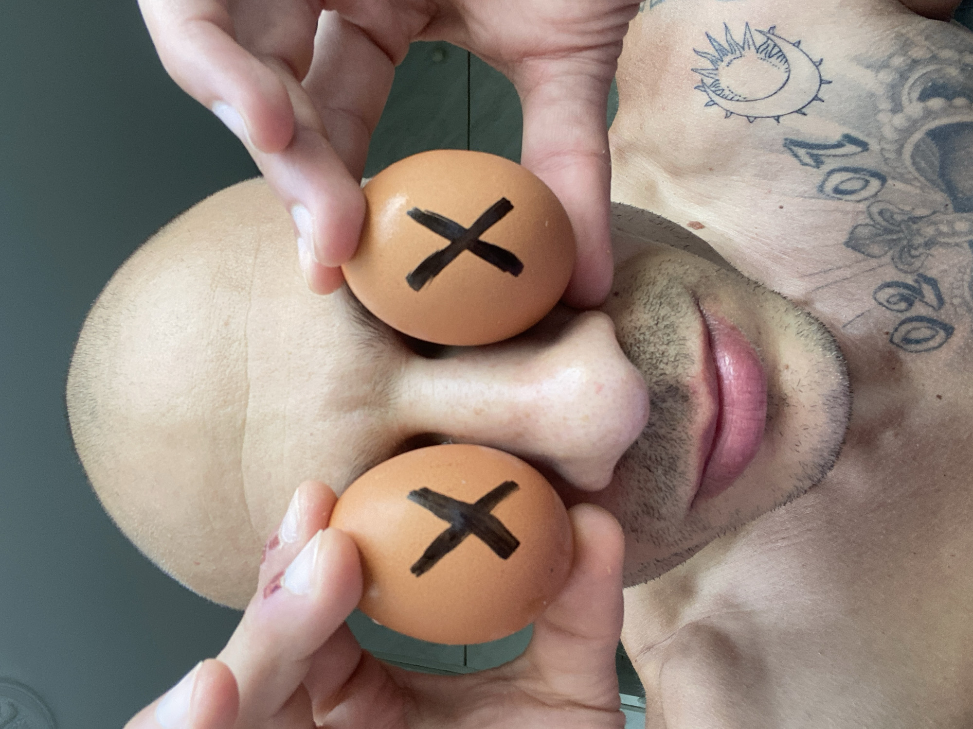 A close-up, front-facing, low-angle shot features a bald person holding two brown eggs, each with a black 'X' drawn on it, up to their closed eyes. The person's hands are positioned to cover their eyes, with the eggs resting against their eyelids. Their gaze is directed upwards, obscured by the eggs. The person's bare shoulders and chest are visible, adorned with tattoos, including a crescent moon with a sunburst and numbers like '02' and '07'. The background is a muted, dark green wall. The image appears to be taken indoors, possibly in a bathroom given the lighting and background elements. There are no discernible connections to the city of Pleven, Bulgaria.