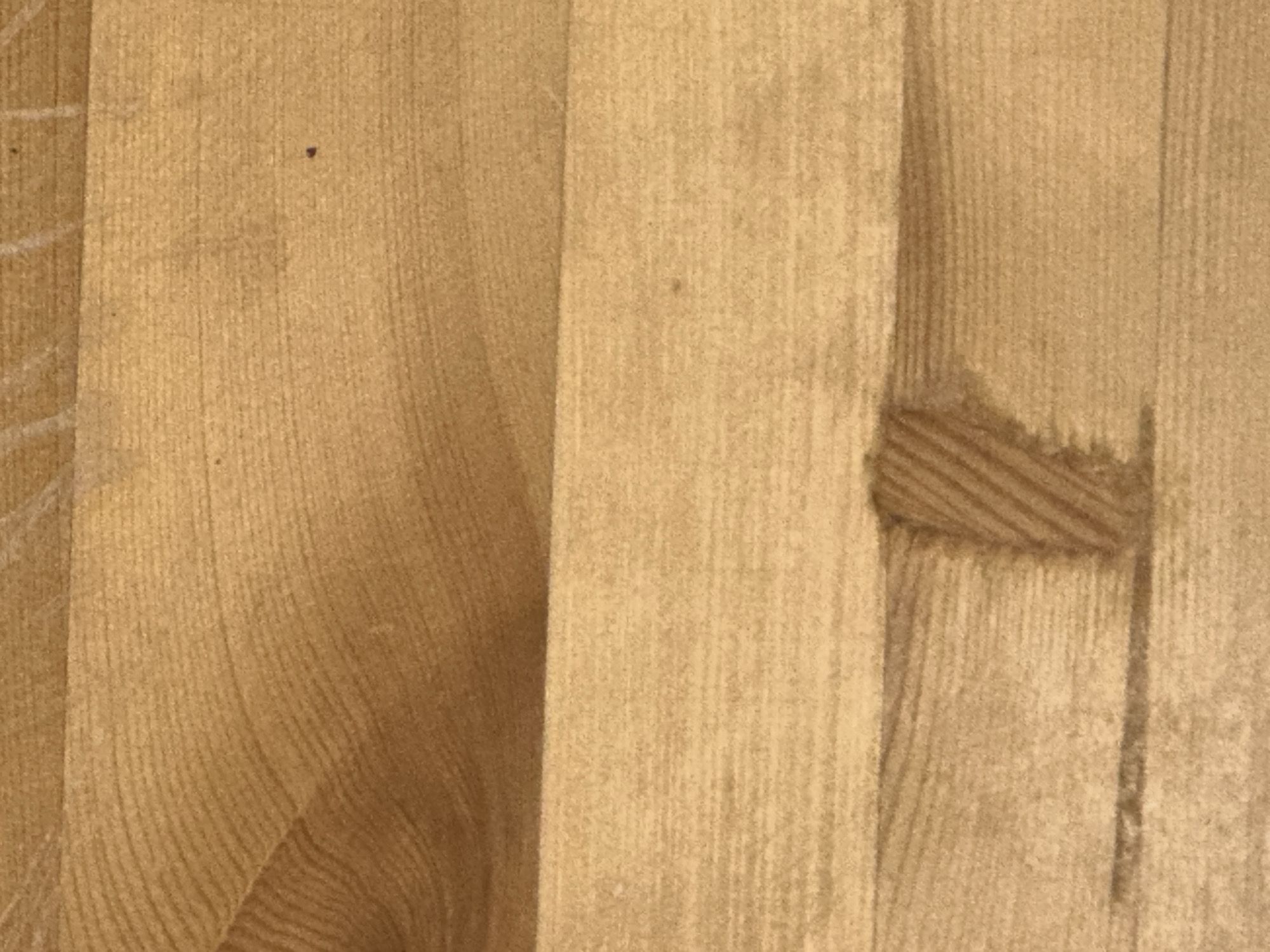 This is a close-up, overhead photograph of a light brown wooden surface. The wood has a visible grain pattern running horizontally across the image, with subtle variations in color and texture. There are faint white lines visible on the upper portion of the surface, possibly from previous markings or wear. In the lower middle section, there is a darker, textured patch that appears to be a knot or a flaw in the wood. The lighting suggests an indoor environment with diffuse illumination, highlighting the natural characteristics of the wood. The image does not provide any contextual information that would allow for its placement within Letterkenny, Ireland, or any specific city.