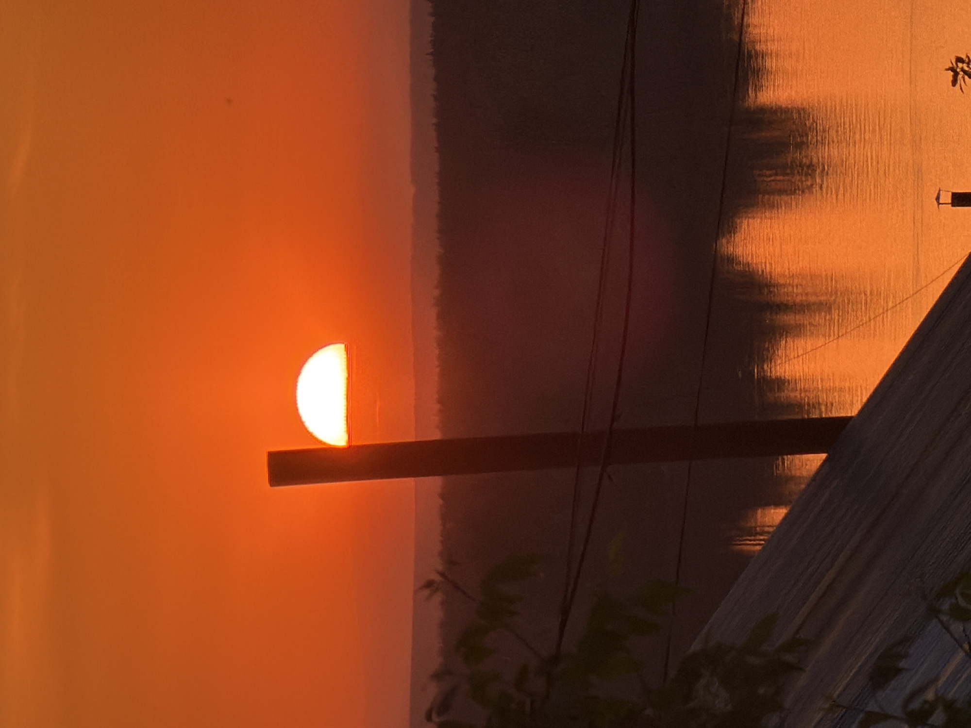 The image depicts a sunset over a body of water, possibly the Kama River, based on the city of Chaikovsky's location in Russia. The sun is partially obscured by a horizontal wooden beam or structure. Power lines are visible in the scene. The overall color palette is orange and warm, characteristic of a sunset. Vegetation is visible in the foreground. The water reflects the light of the setting sun. The other side of the body of water appears to be a forest or land area. A portion of a structure, possibly a building or dock, is visible in the lower right corner.