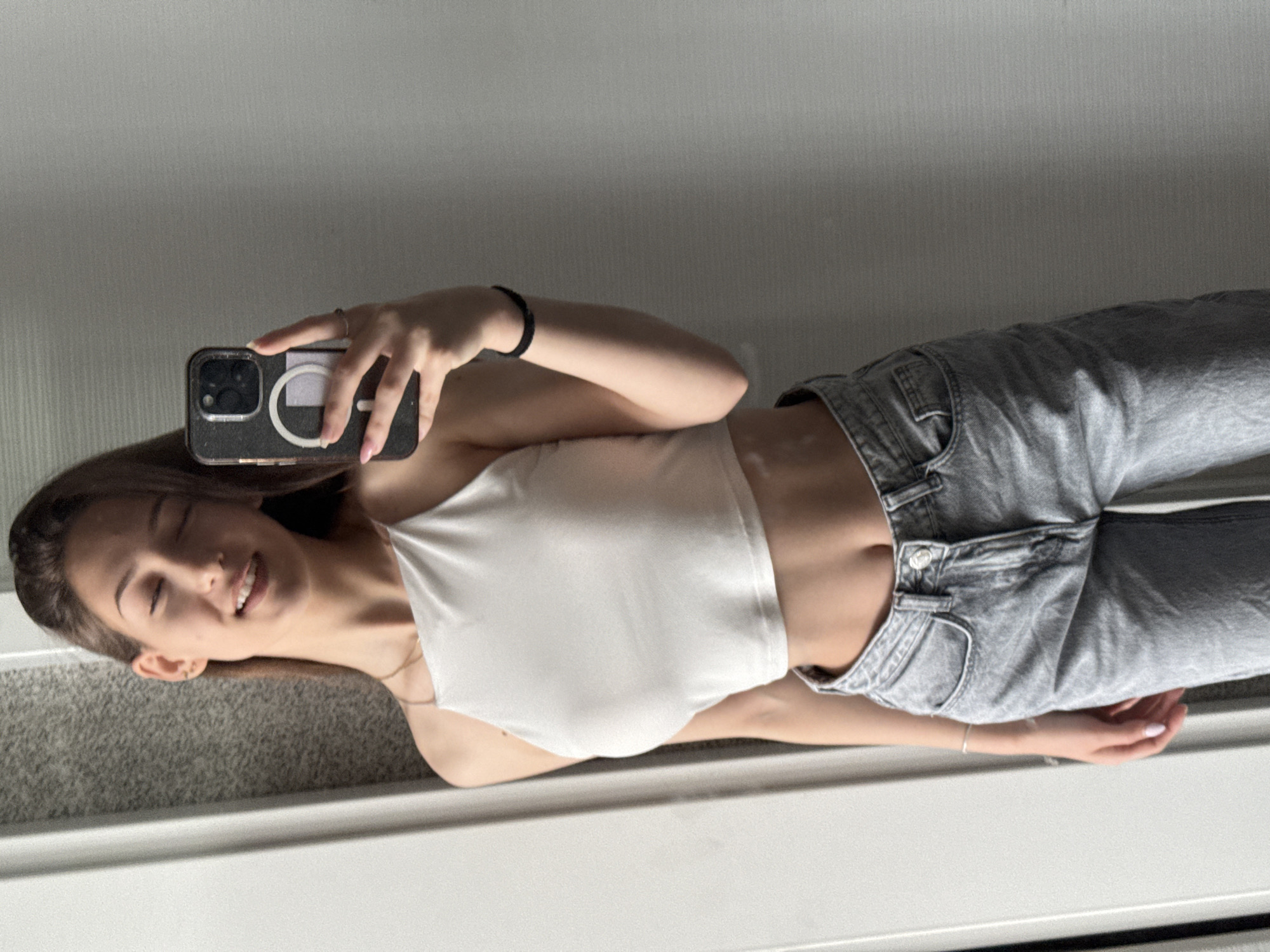 A young woman is taking a selfie in front of a mirror. She is wearing a white crop top and light-wash jeans. She is holding a smartphone with a dark-colored case with a white circular magnet on the back. The background appears to be a light-colored wall. The scene's location in Sofia, Bulgaria, cannot be determined from the visual information provided.