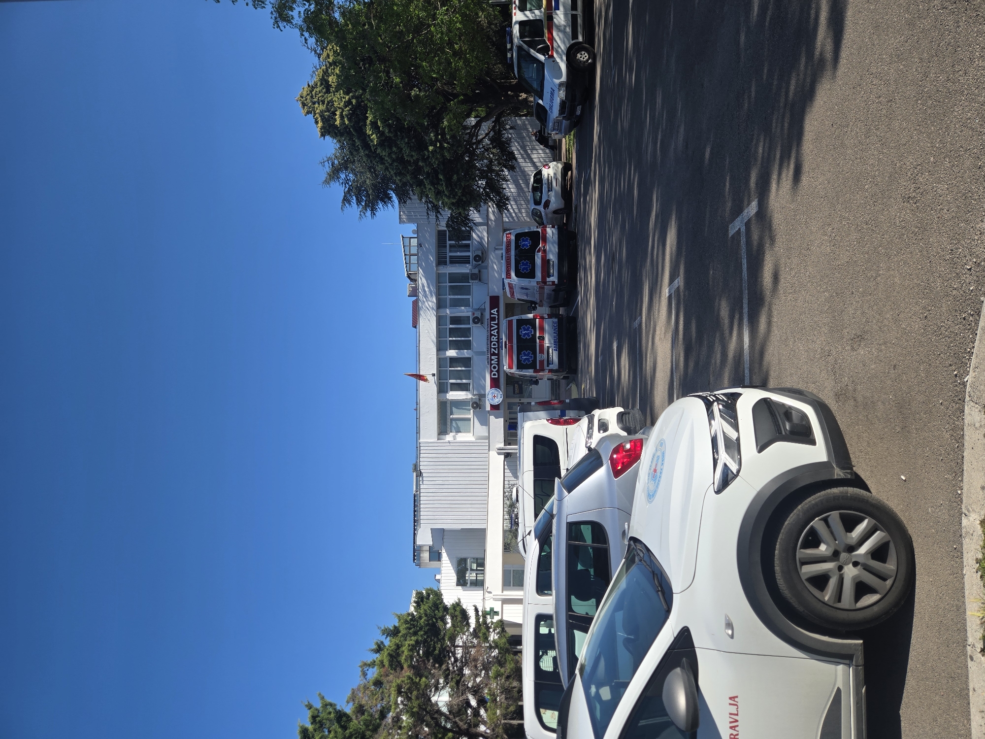 The image shows a street scene with a row of parked vehicles in front of a white building identified as "DOM ZDRAVLJA." Several of the vehicles are white vans and ambulances. The location is not recognizable based on the provided information. The sky is clear and blue. Trees are present on either side of the building, casting shadows on the parking area. The scene appears to be outdoors during daylight.