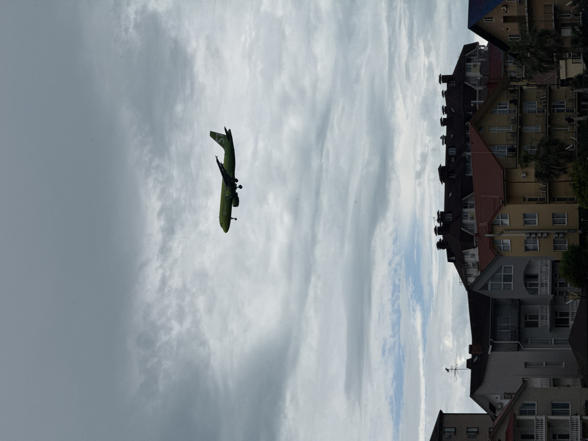 A low-angle, overcast shot shows an S7 Airlines aircraft on approach for landing, with the skyline of a city – likely Sochi, Russia – as a backdrop. The aircraft is lime green and flying toward the photographer. The multi-story buildings are varying shades of white, beige, and gray and are topped with dark roofs. Tall, narrow clouds are visible beyond the airplane.