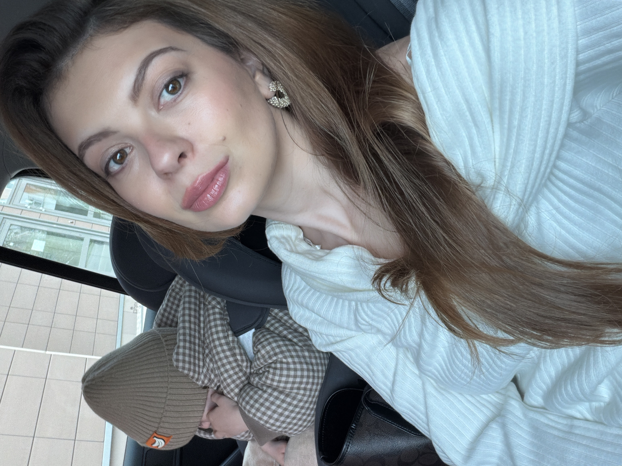 A light-skinned woman with brown hair and brown eyes is in a car. She is wearing a white, ribbed sweater and a gold earring. A child is wearing a brown plaid shirt and a brown beanie. The picture was taken in Khimki, Russia, as can be seen through the window.