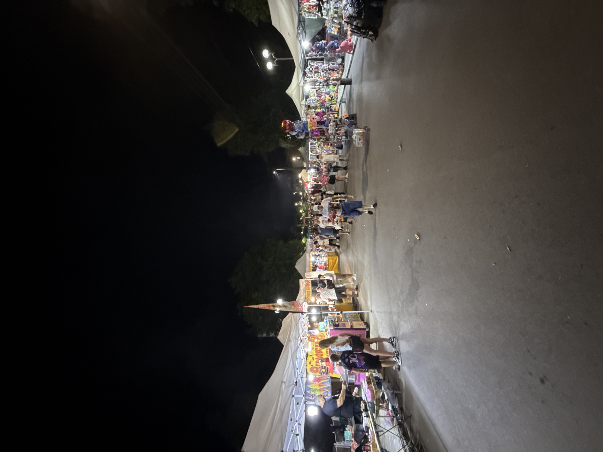 The image depicts a night scene at an outdoor market or fair, likely in the city of Stob, Bulgaria. The scene is illuminated by artificial lights from the stalls and streetlights, creating a lively atmosphere. The market extends along a paved walkway, with numerous tents and stalls set up on either side. These stalls appear to be selling a variety of goods, including food items like ice cream and cotton candy, as well as toys and souvenirs.
Several people are visible, walking along the path, browsing the stalls, and interacting with vendors. Some are dressed casually, indicating a relaxed evening outing. The overall impression is one of a community gathering or a popular tourist attraction. The background shows trees and the dark night sky, suggesting an urban or semi-urban setting. The ground surface is asphalt or a similar paved material.