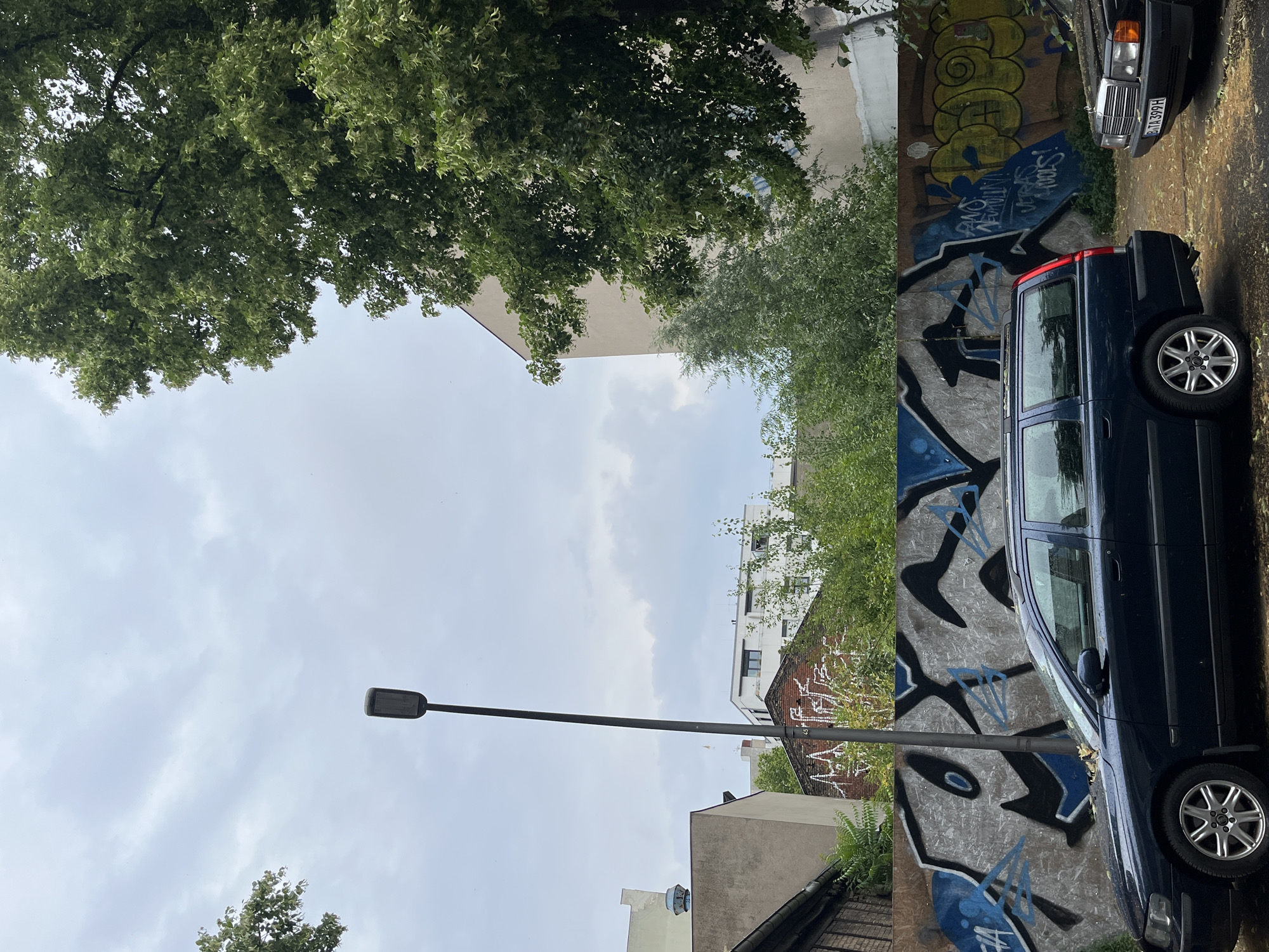 A dark blue Volvo station wagon is parked in front of a graffiti-covered wall in Berlin, Germany. The graffiti is abstract and features blue and black spray paint. A second, older black Mercedes-Benz sedan is parked to the right of the Volvo. A tall, dark grey streetlamp stands to the left of the graffiti wall. Behind the wall, trees and buildings are visible. The sky is overcast.