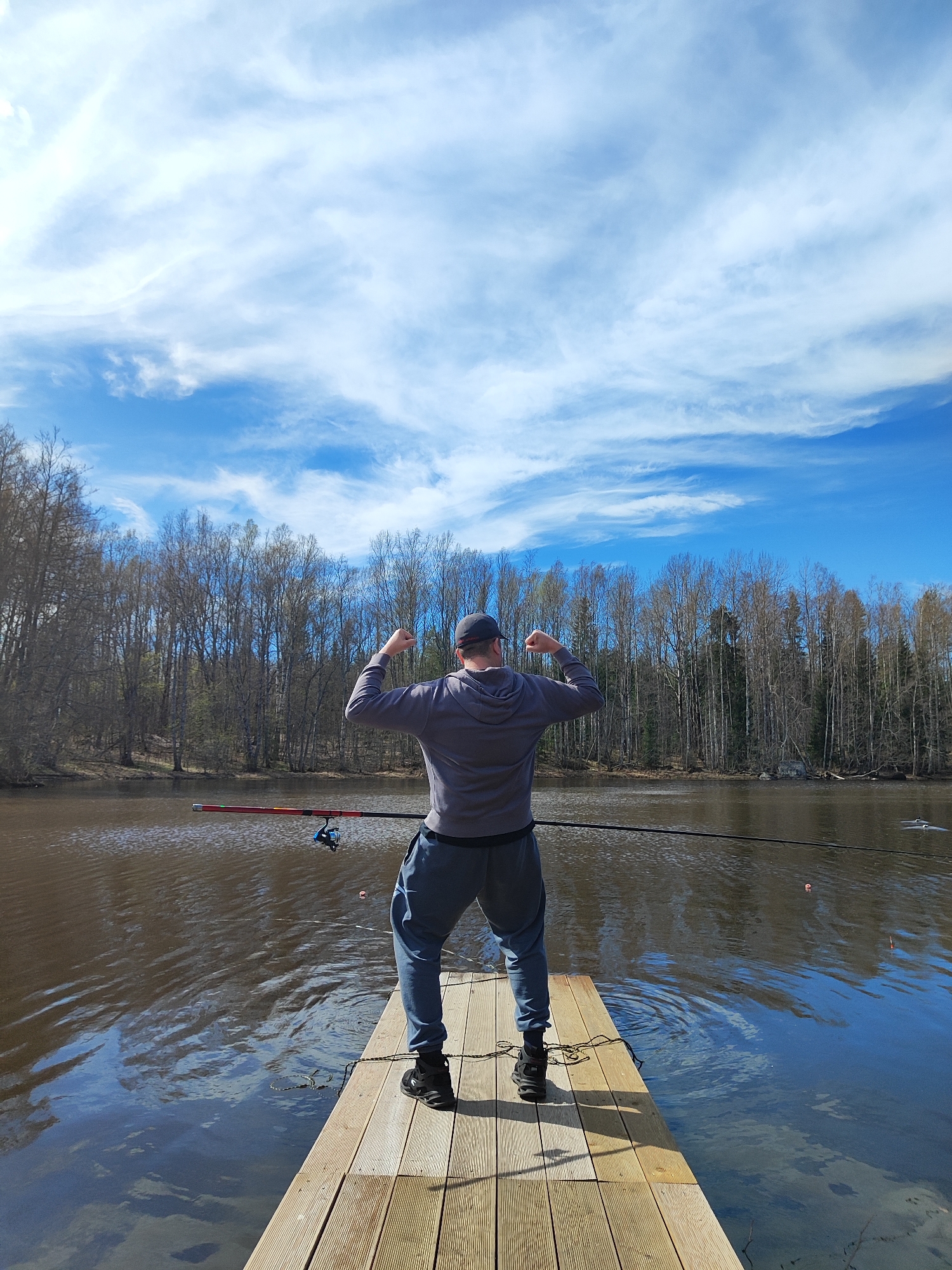A man stands on a wooden dock at the edge of a body of water. He is seen from the back, wearing a grey hooded sweatshirt, grey sweatpants, a dark cap, and black shoes. His arms are bent, fists clenched, and raised towards his shoulders in a flexing gesture. A fishing rod rests on the dock behind him, its line extending into the water. Behind the dock, across the water, is a line of bare trees. Above the trees is a blue sky with white clouds. The location appears to be in a natural setting, but the city remains unknown.