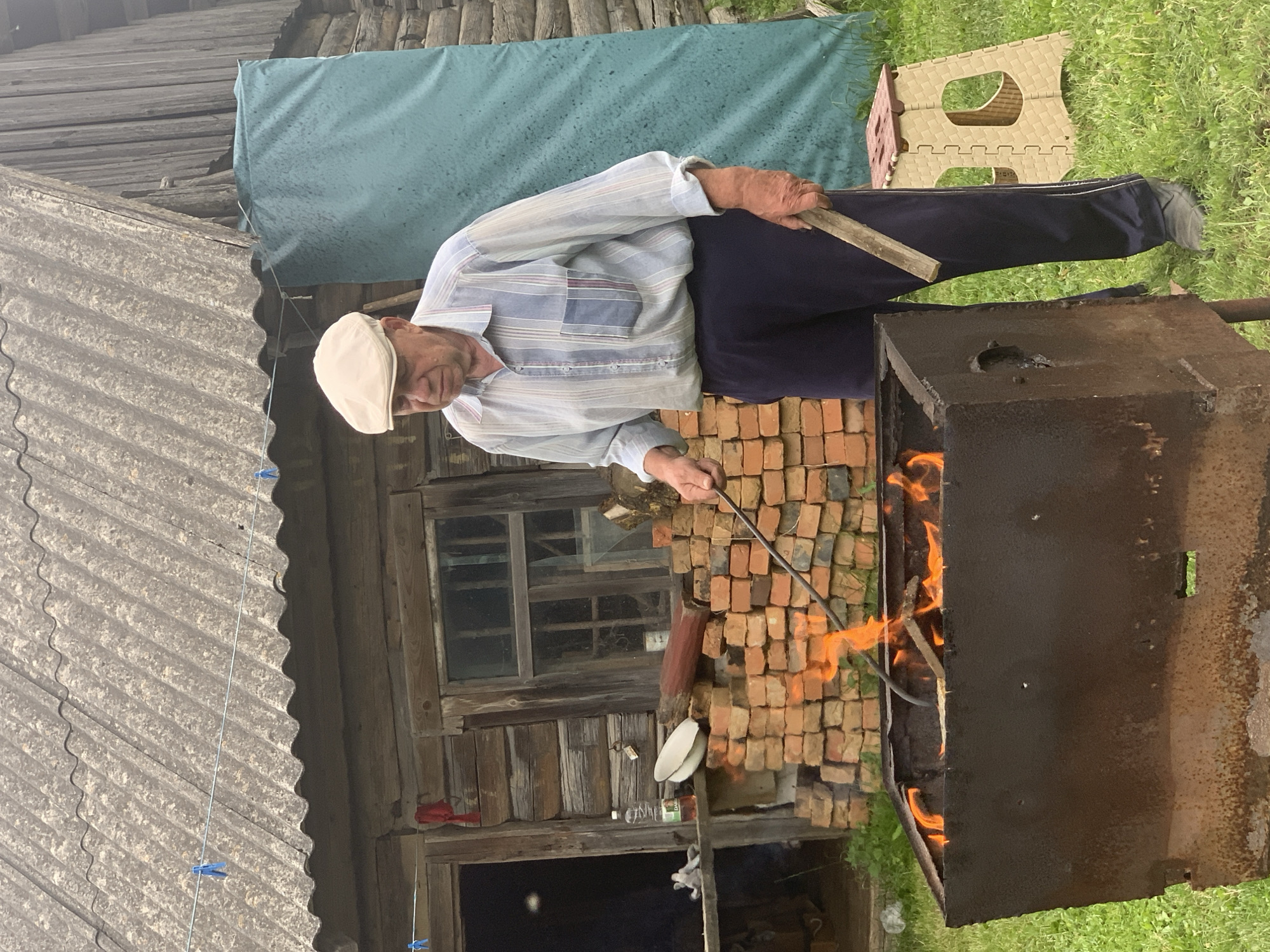 An elderly man wearing a light-colored cap, a striped button-down shirt, and dark pants stands in front of a rusty barbecue grill with flames. He holds a metal skewer in his right hand and a wooden plank in his left. The background features a rustic wooden building with a corrugated roof, a stack of bricks, and a large green tarp. The scene appears to be outdoors in a rural or semi-rural setting, possibly in the Відзаўскі сельскі Савет, Belarus. The ground is covered in green grass. A small, collapsible stool is visible to the right of the man. Clotheslines with blue clothespins are strung across the roof of the building.