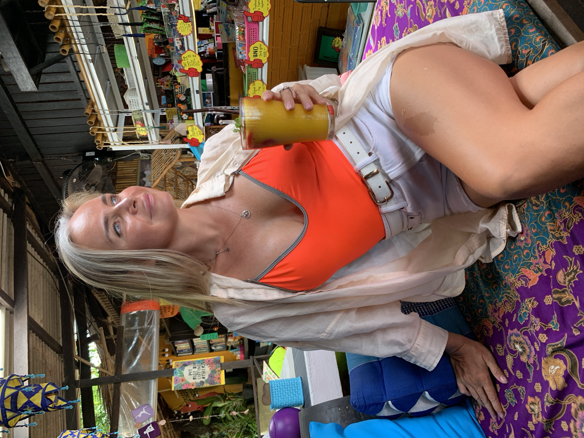 A blonde woman is sitting on a bench with a purple floral patterned cushion. She is wearing an orange bikini top, white shorts, a light beige button-up shirt, and a white belt. She is holding a tall glass of a yellow-orange beverage in her right hand. Her left hand is resting on the cushion. She is looking to her right. Behind her, there are shelves filled with various items, including books, trinkets, and colorful signs with Thai writing. A fan is visible on the left side of the image. The setting appears to be an outdoor market or cafe in Baan Bo Phut, Thailand, indicated by the style of the building, the merchandise on the shelves, and the overall tropical ambiance.