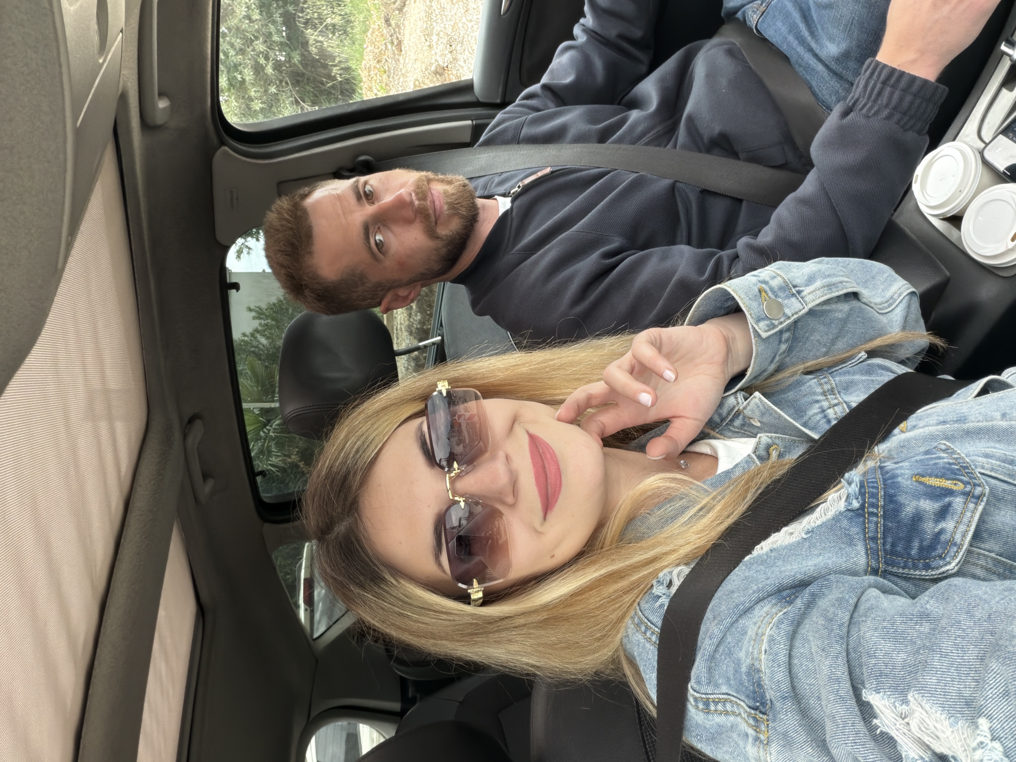 A medium-shot, canted selfie shows a man and woman inside a car. The woman is in the foreground, wearing sunglasses and a denim jacket, with her hand near her face. The man is behind her and to the side, with a beard and wearing a dark sweatshirt and jeans. Both are wearing seatbelts. There are two sealed coffee cups visible in the center console. The view outside the car window is out of focus but suggests vegetation, potentially near Lastva Grbaljska, Montenegro.