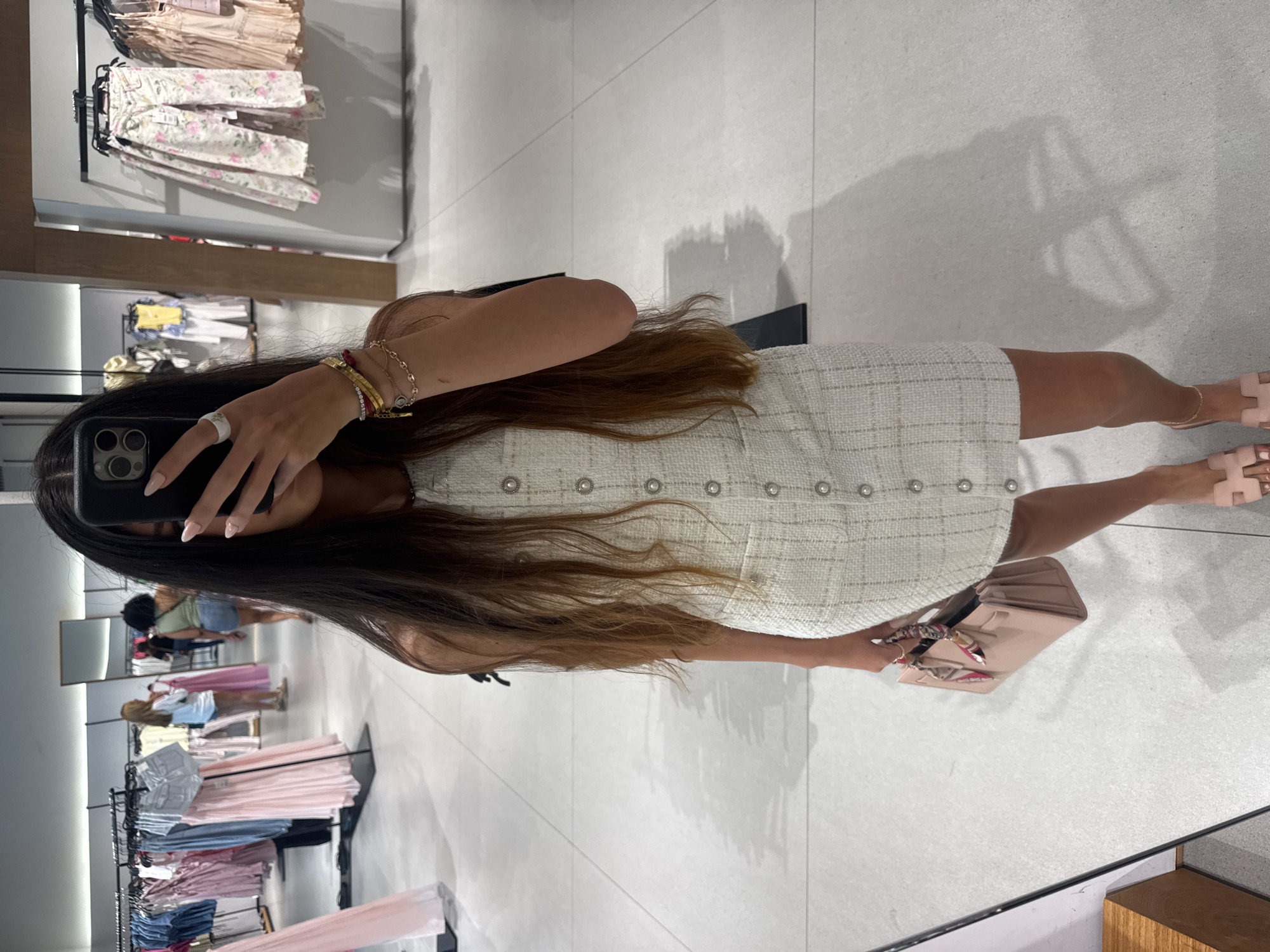 A full-body, waist-up mirror selfie of a woman in a white tweed dress with pearl buttons, holding a pale pink handbag with a colorful scarf tied to the handle. She is wearing delicate gold bracelets and a ring on her right hand, which is partially obscuring her face as she holds her phone to take the photo. Her long, dark hair cascades down her back. She is also wearing pale pink flat sandals. The background shows a clothing store with racks of clothes, including pink and blue dresses and floral-patterned pants. The floor is tiled with large, light-colored tiles, and shadows are cast on the floor. The setting appears to be a brightly lit retail environment.