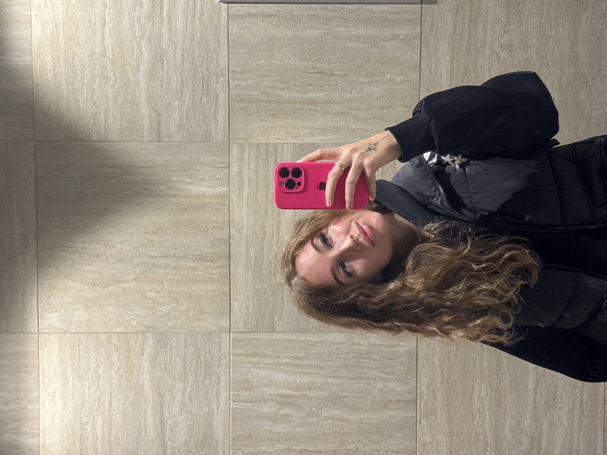 A young woman is taking a selfie in a bathroom or restroom with beige tile walls. She is holding a bright pink iPhone in her right hand. She has long, wavy brown hair and is wearing a black puffer vest and a black top with long sleeves. There is a small tattoo on her left hand. The location could be in Сокол, Russia.