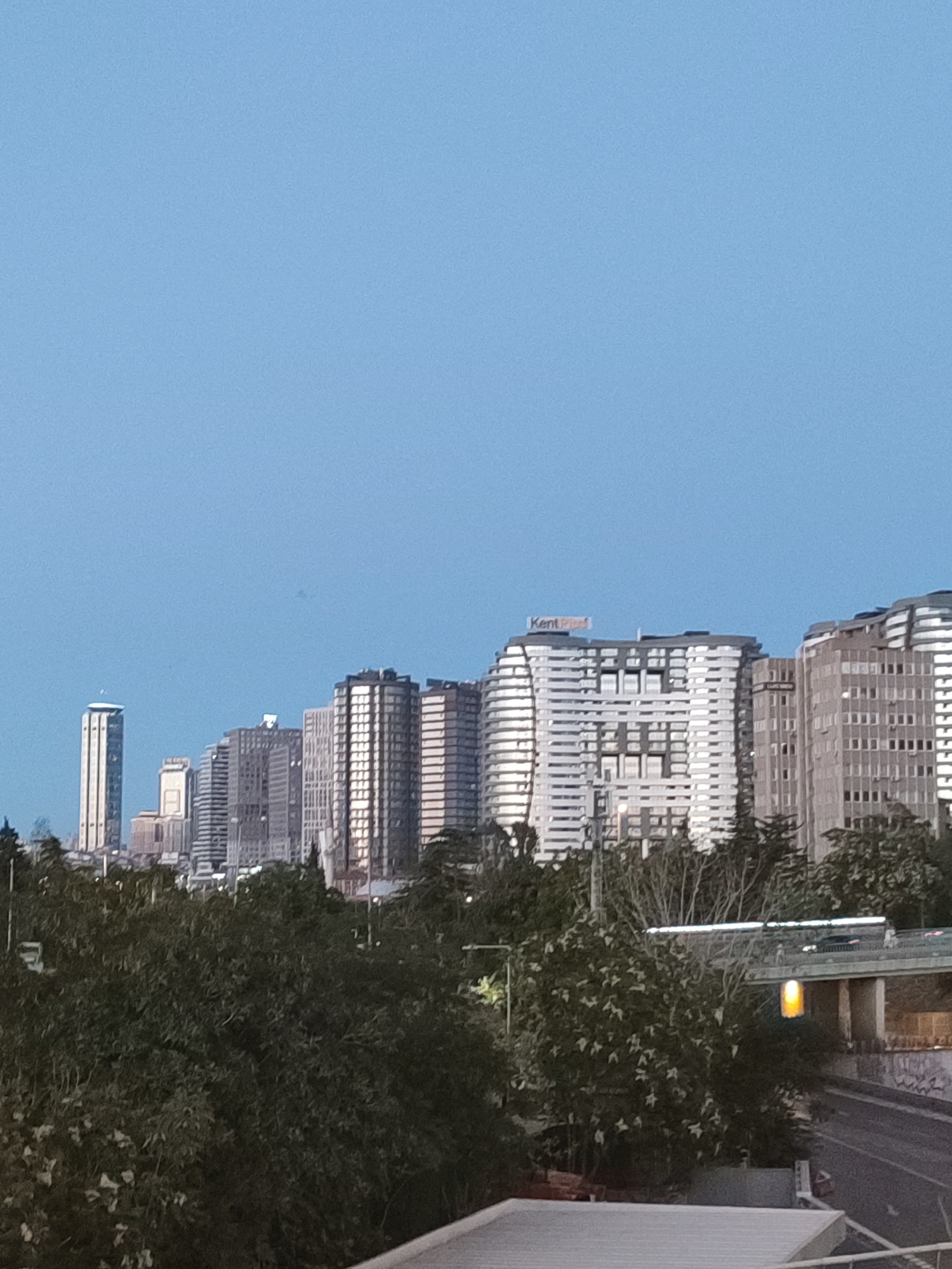 The image shows a panoramic view of a city skyline with numerous tall buildings. The sky is clear and a pale blue, suggesting it is either early morning or late evening. In the foreground, there is a dense canopy of dark green trees, partially obscuring the lower parts of the buildings.
The buildings vary in height and architectural style. Some are sleek and modern, with glass facades, while others appear older, with more traditional concrete and brick structures. One particularly tall, slender skyscraper stands out on the left side of the frame, topped with a distinctive circular structure. The buildings in the center and right appear to be clustered together, forming a dense urban landscape. A sign with the word "KentPlus" is visible on the rooftop of one of the buildings.
Below the main skyline, a road and what appears to be an overpass are visible, suggesting a busy transportation network. A bright yellow light hangs from a pole over the road, illuminating the area. There are no people or vehicles clearly visible in the image. The overall impression is of a modern, bustling city seen from a distance, with nature playing a supporting role in the foreground.
The image is consistent with a view of the Kadıköy district in Istanbul, Turkey, known for its mix of residential and commercial high-rises.