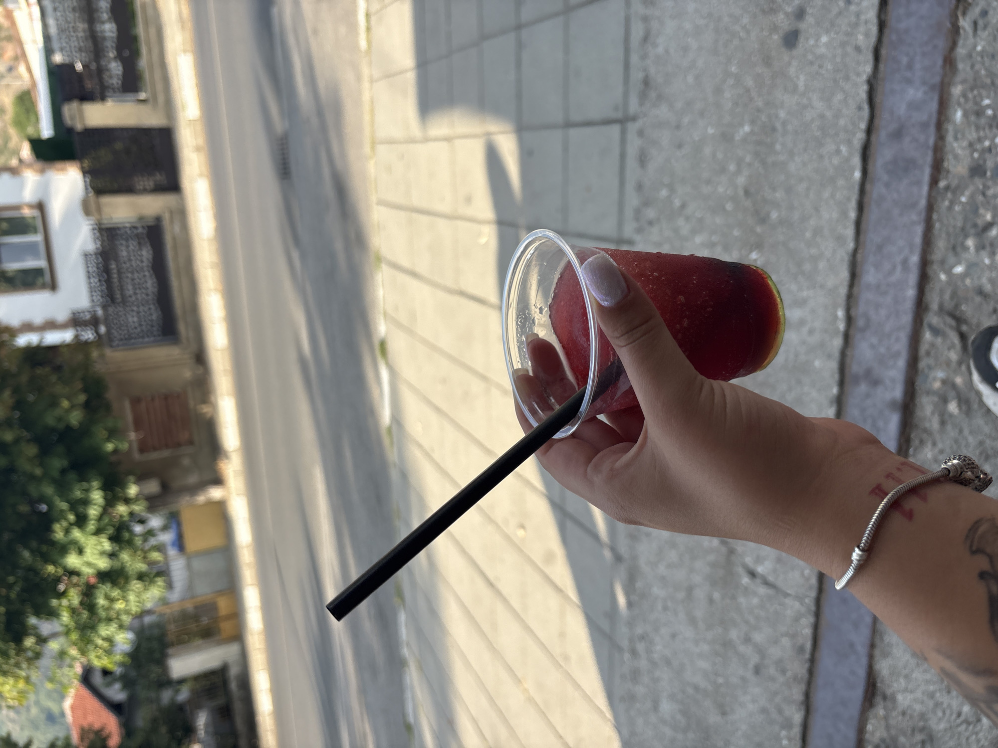 A close-up shot captures a person's hand holding a clear plastic cup filled with a red, frozen beverage and a black straw. The cup is condensation-covered, indicating it contains a cold drink. The hand, adorned with a silver bracelet and a tattoo on the wrist, gently grasps the cup. In the background, a street scene unfolds, featuring a paved sidewalk, a road, and buildings with architectural details characteristic of Sopot, Bulgaria. The sunlight casts shadows across the sidewalk, suggesting a daytime setting. The overall ambiance is relaxed and casual, hinting at a moment of enjoyment on a warm day.