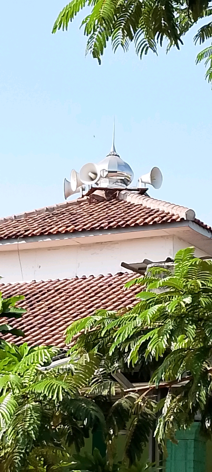 This is a photograph of the roof of a building in Satriamekar, Indonesia. The roof is covered with reddish-brown tiles and has a white wall below it. Atop the roof is a silver, dome-shaped structure with a pointed finial. Attached to the dome are at least three white loudspeakers. Green foliage from trees overhangs the roof from the top and bottom of the frame. The sky is clear and blue.