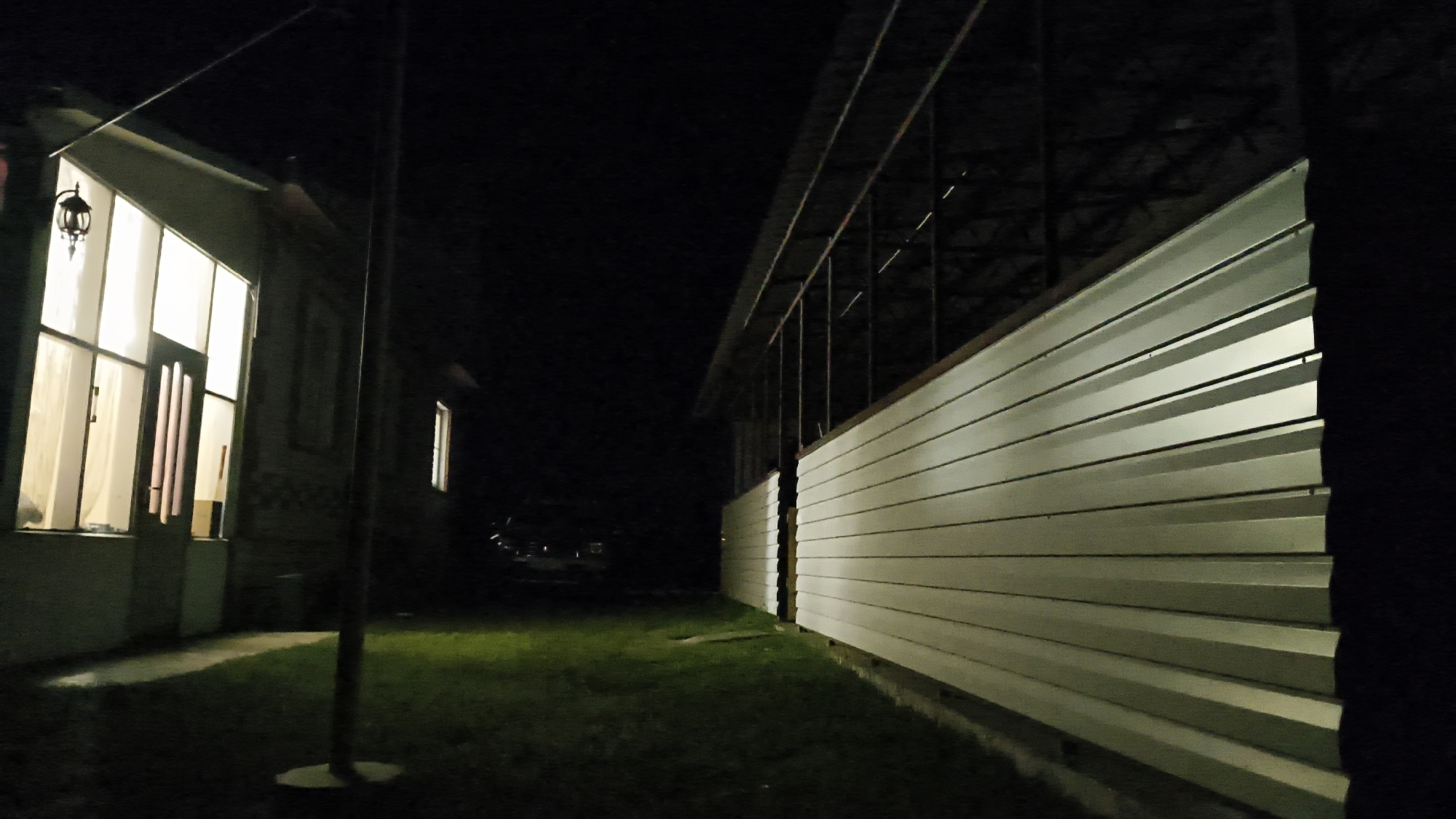 The image depicts a nighttime scene in Sharashenskii, Russia. On the left, a building with large, illuminated windows and a hanging lantern is visible. A dark pole stands in the foreground between the building and a long, horizontal structure on the right. This structure is covered with light-colored, horizontal siding and extends into the background. The ground between the two structures is covered in grass. In the background, a dimly lit car is parked. The overall scene suggests a residential or commercial area at night.