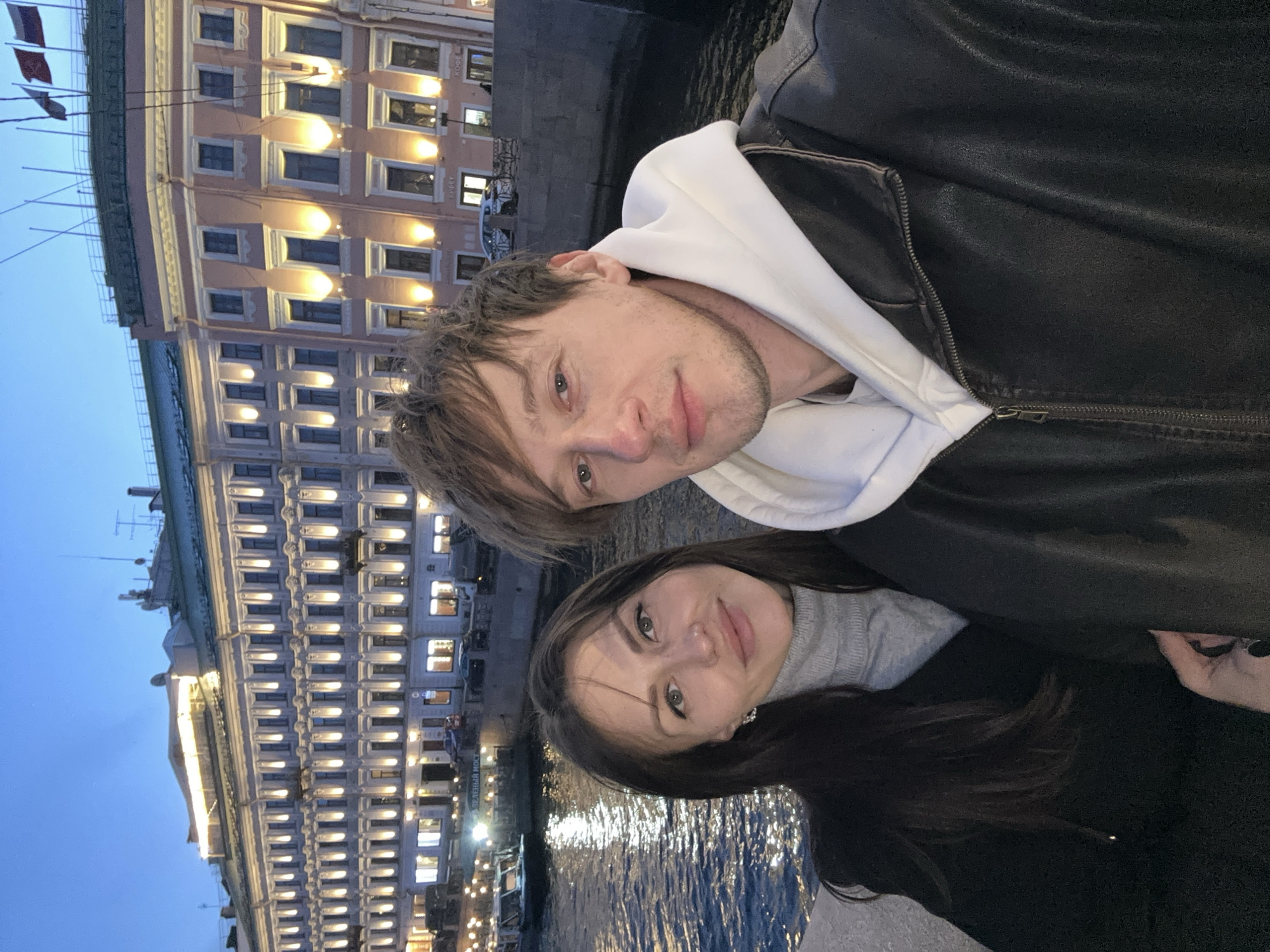 Two people, a man and a woman, are in a close embrace, posing for a selfie, against the backdrop of a city landscape at dusk. The man, in the upper portion of the frame, has fair skin and short, light brown hair. He is wearing a white hooded sweatshirt underneath a black leather jacket. His gaze is directed towards the camera. Below him and slightly to the left, the woman has dark hair and is wearing a grey sweater. She is also looking towards the camera. The background features a large, ornate building illuminated with numerous windows, reflecting the soft, evening light. A body of water, likely a river or canal, can be seen in the lower portion of the frame. The building's architectural style and the presence of water suggest this photograph was taken in Saint Petersburg, Russia. A bridge, Zeleniy Most, is partially visible.