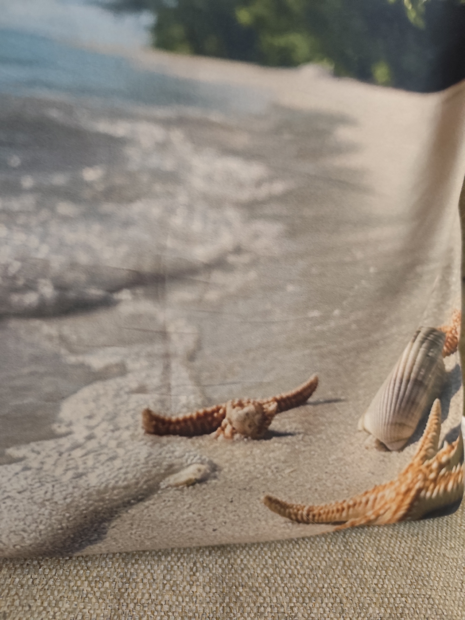 The image shows a close-up of a textured fabric, possibly a cushion or upholstery, with a printed design of a beach scene. The design features a sandy beach with gentle waves lapping at the shore. Two starfish and a seashell are depicted on the sand. One starfish is positioned in the lower left, partially obscured by the fabric's texture. Another starfish is in the lower right, with its arms spread. A ridged seashell is located between the two starfish. The background of the design shows blurred green foliage and the ocean. The fabric itself has a visible weave, appearing to be a neutral beige or light brown color. There are no people or animals present in the image. The location of Montana, Bulgaria is not depicted in the image, as the scene is a generic beach setting.