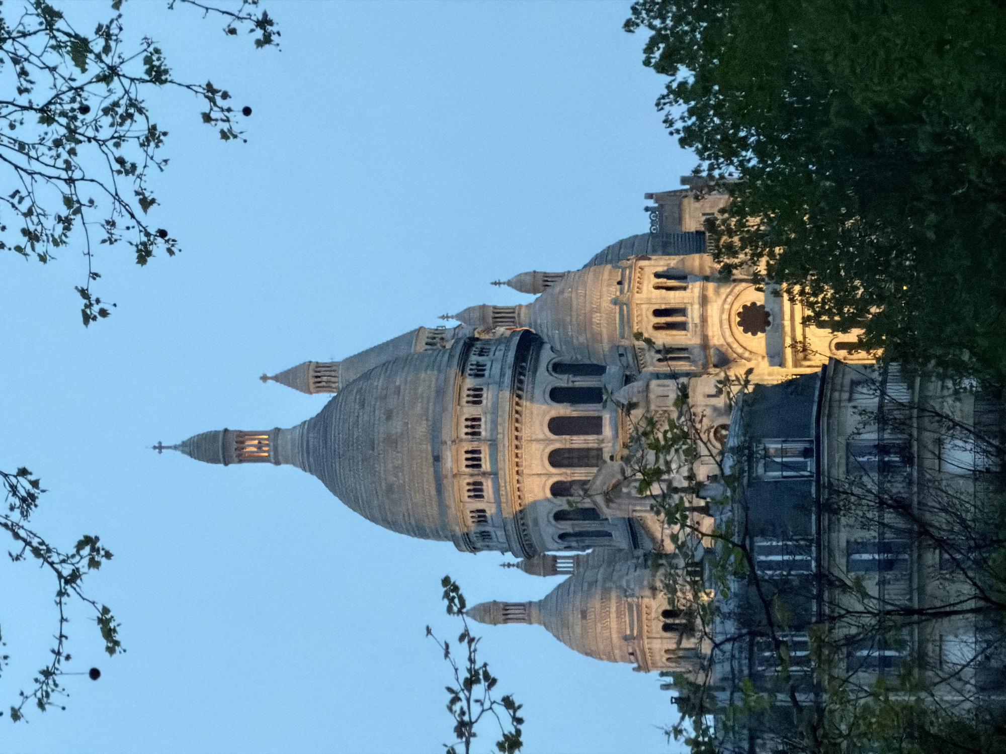 The image features the Sacré-Cœur Basilica in Paris, France, framed by foliage in the foreground. The basilica's domes and spires are illuminated against a blue sky. Tree branches with sparse leaves are visible on the left, while dense foliage is on the right side of the frame. The building's architectural details are visible, including windows, arches, and decorative elements. The image is taken from a low angle, looking up at the basilica.